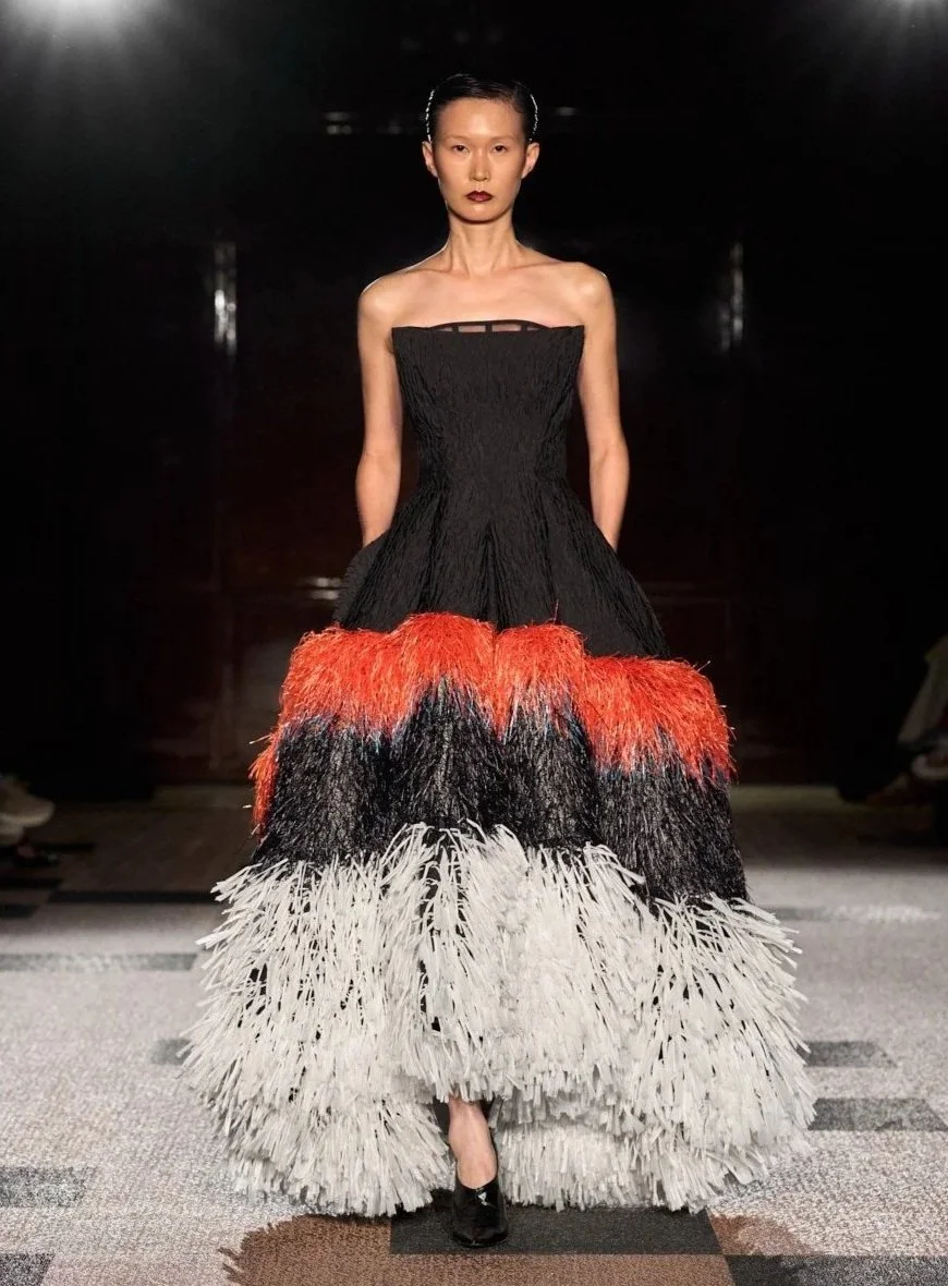 A model walking on a runway wearing a strapless black dress with a colorful feathered skirt in white, black, orange, and red, with a dark background.