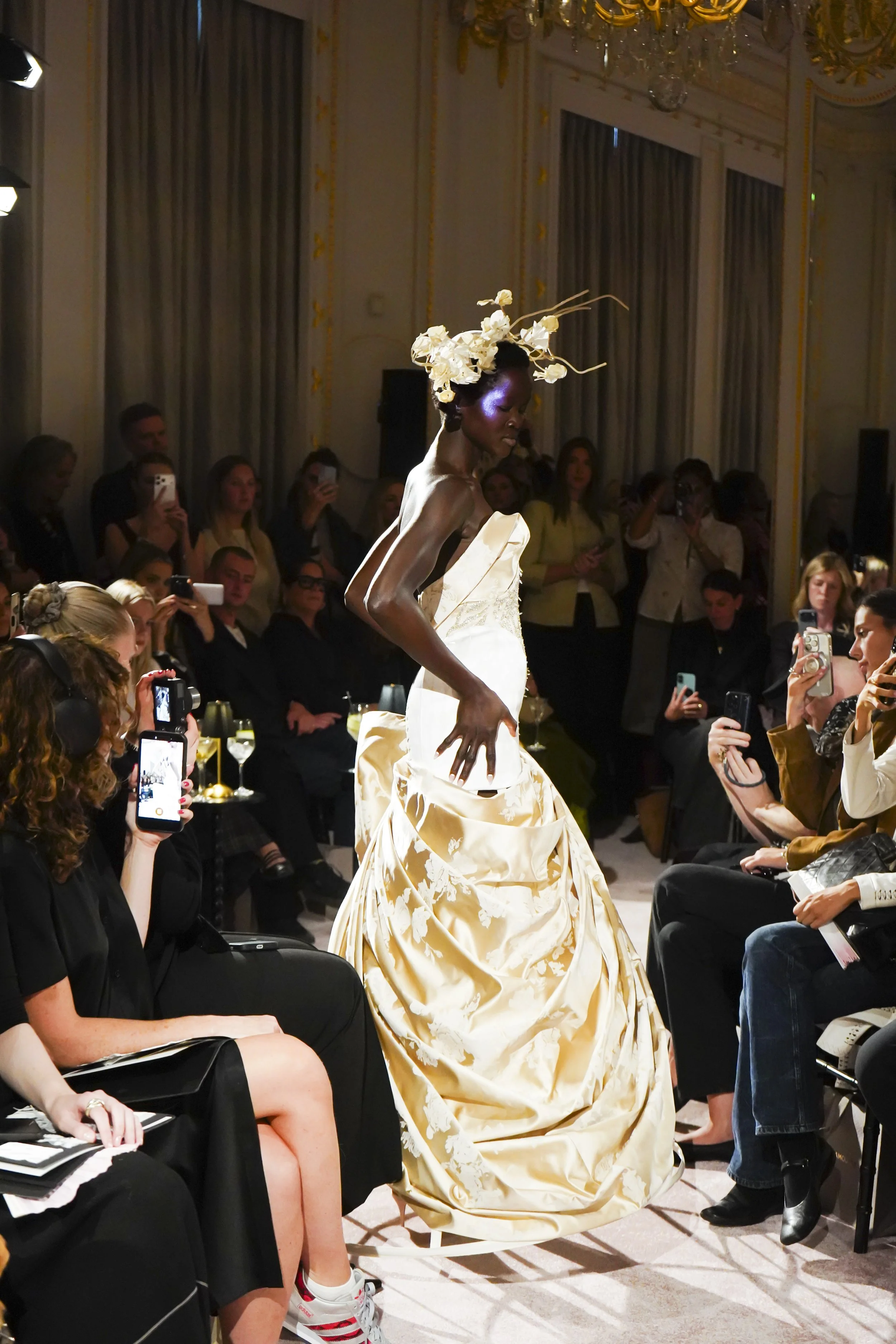 A model in a sculptural ivory satin gown and floral headpiece walks past the front row.