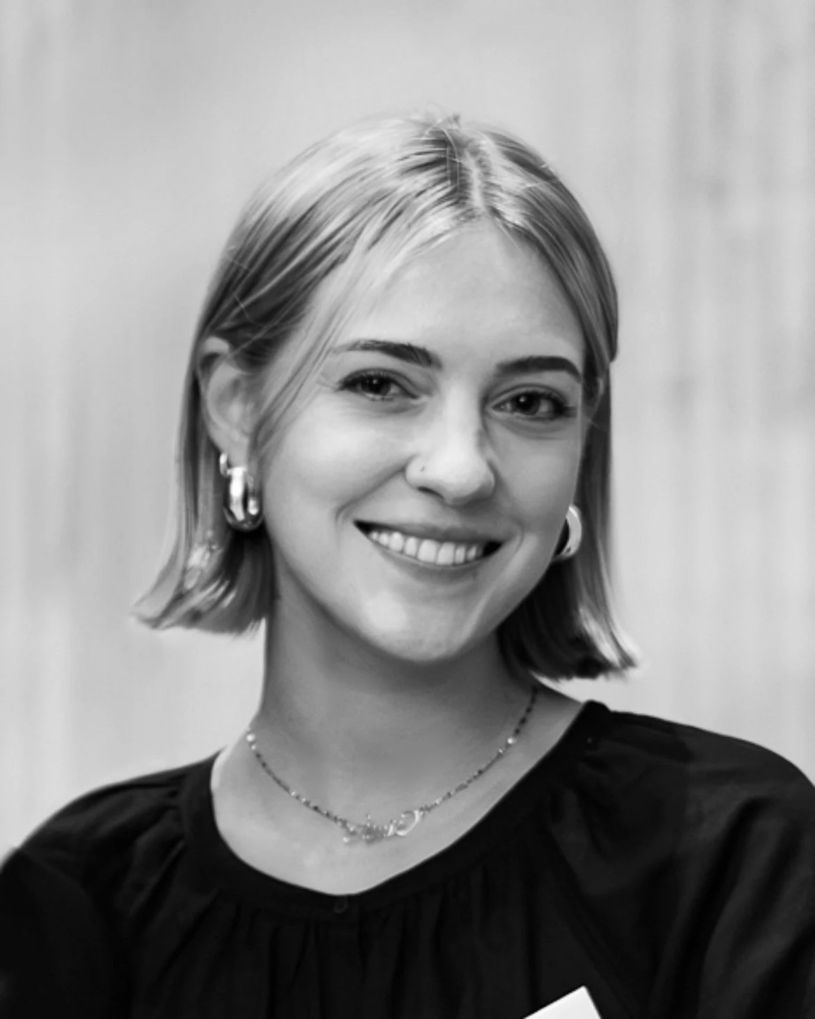 Black and white portrait of a young woman smiling, with shoulder-length hair, hoop earrings, a nose piercing, a necklace, and a dark top.
