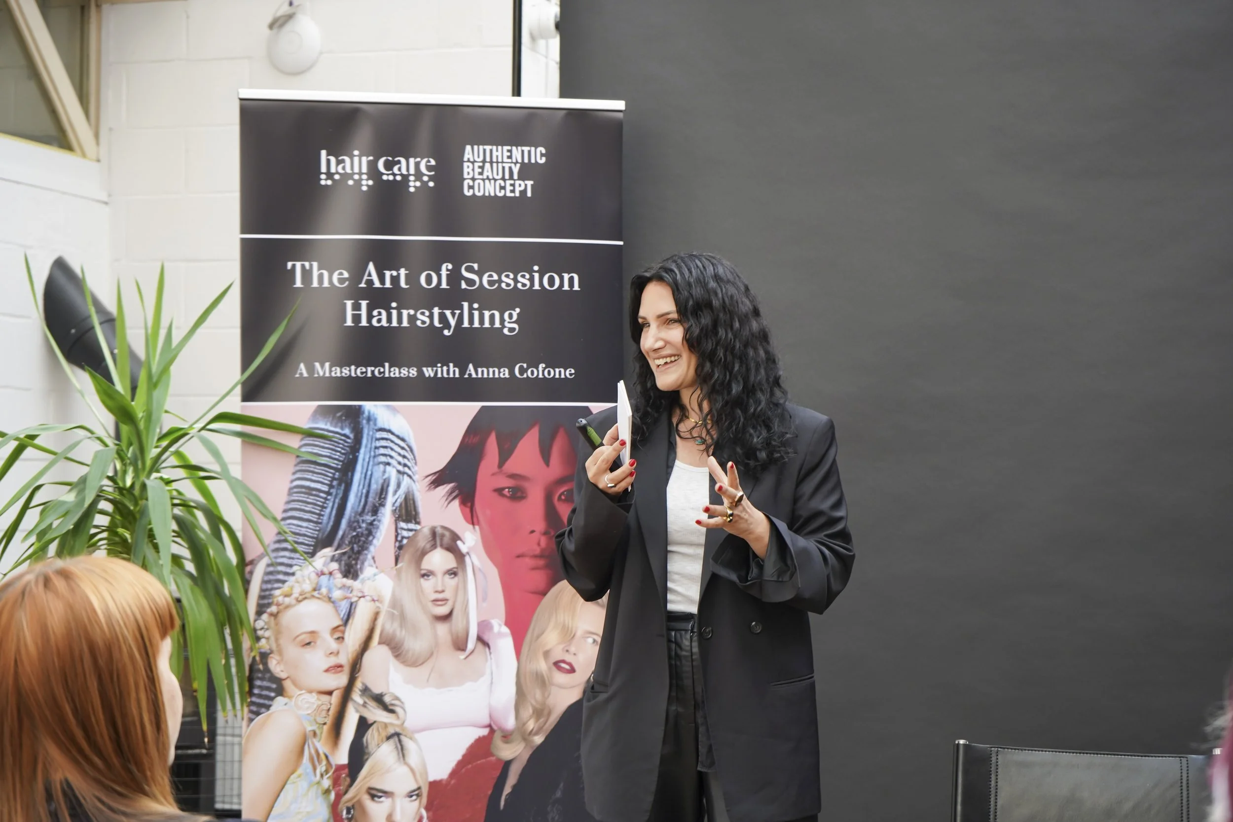 A woman with curly dark hair and a black blazer speaking during a hair care masterclass, against a black background and banner