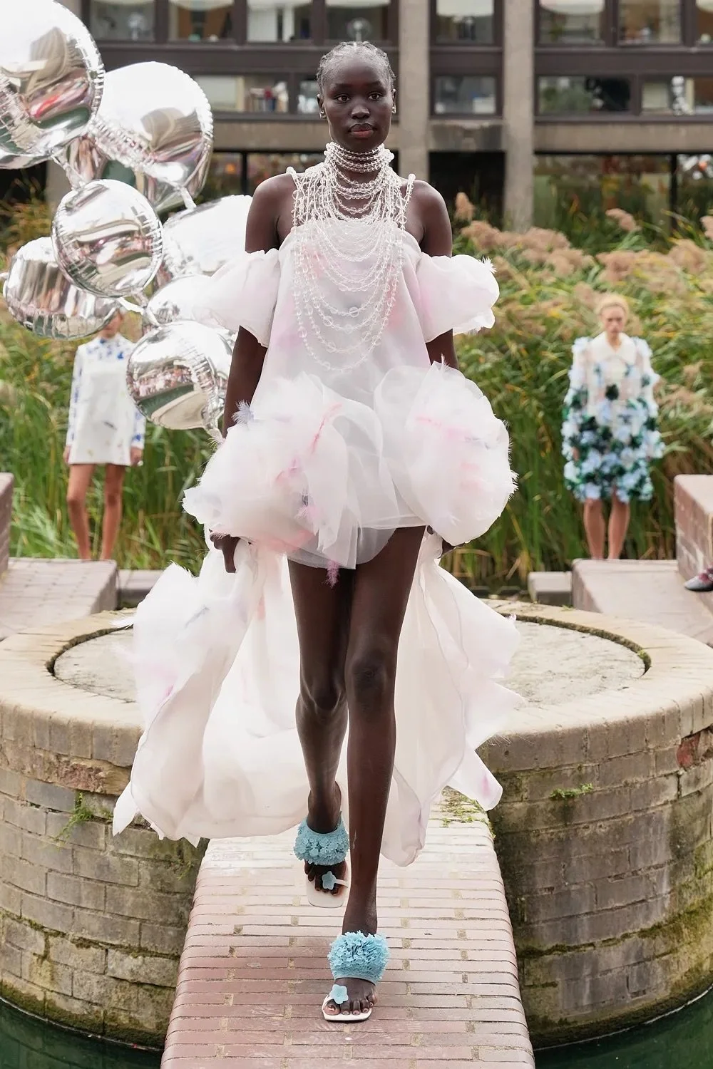 A model in a sheer white dress with puffed sleeves and layered beaded necklaces walks a brick catwalk.