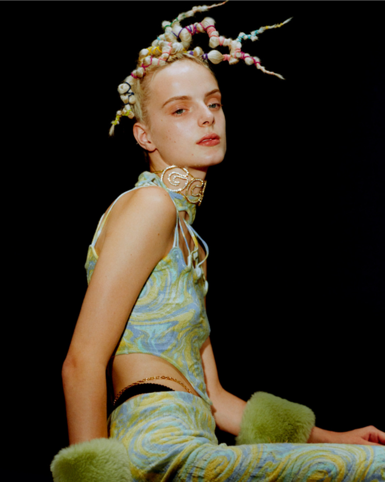 A fashion shot of a model with colourful, curled hair styled in dreadlocks posing against a black background, wearing a patterned satin top with fuzzy green wrist cuffs and a necklace.