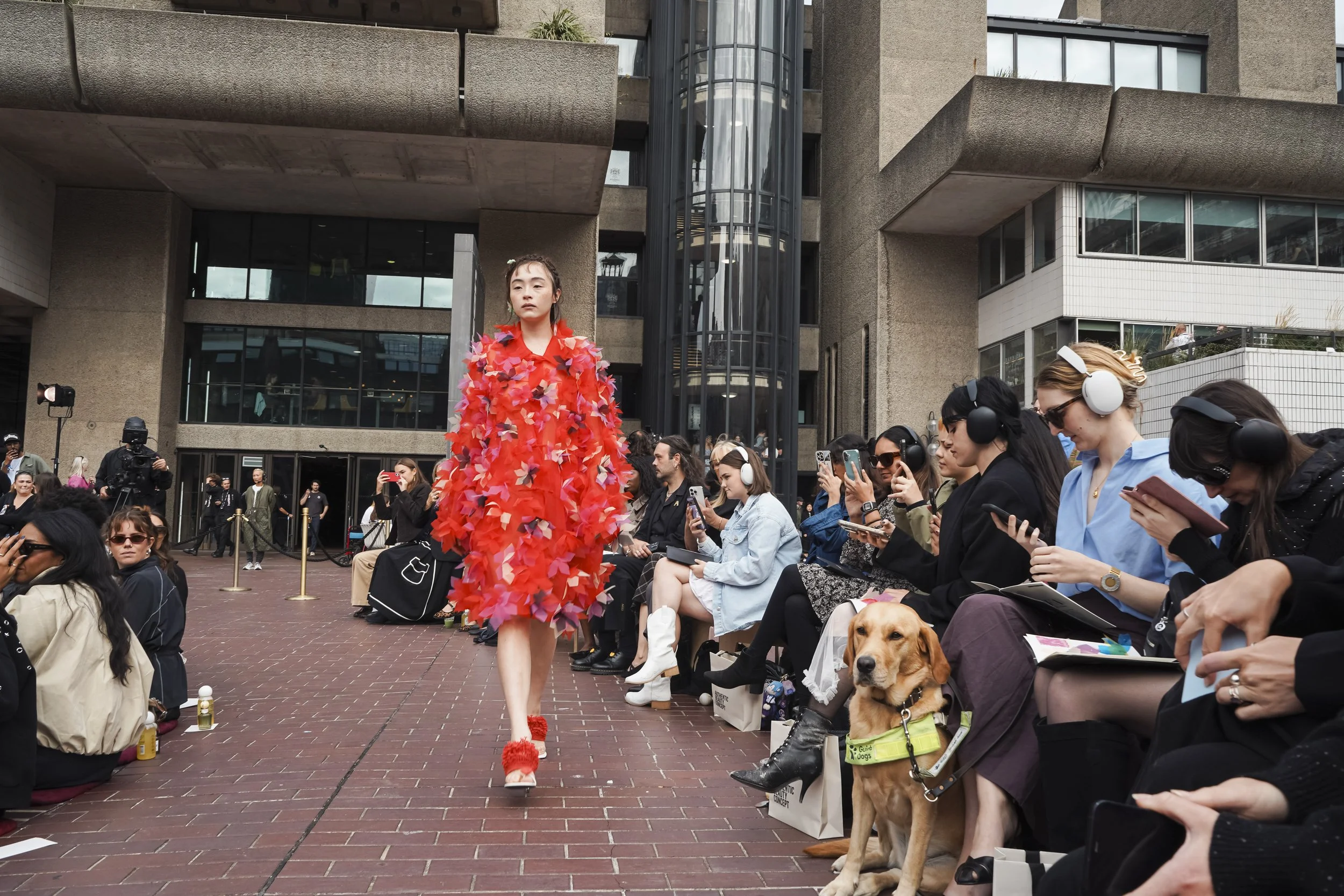 A model in a red coat covered in 3D petals walks an outdoor catwalk past guests wearing headphones and a guide dog.