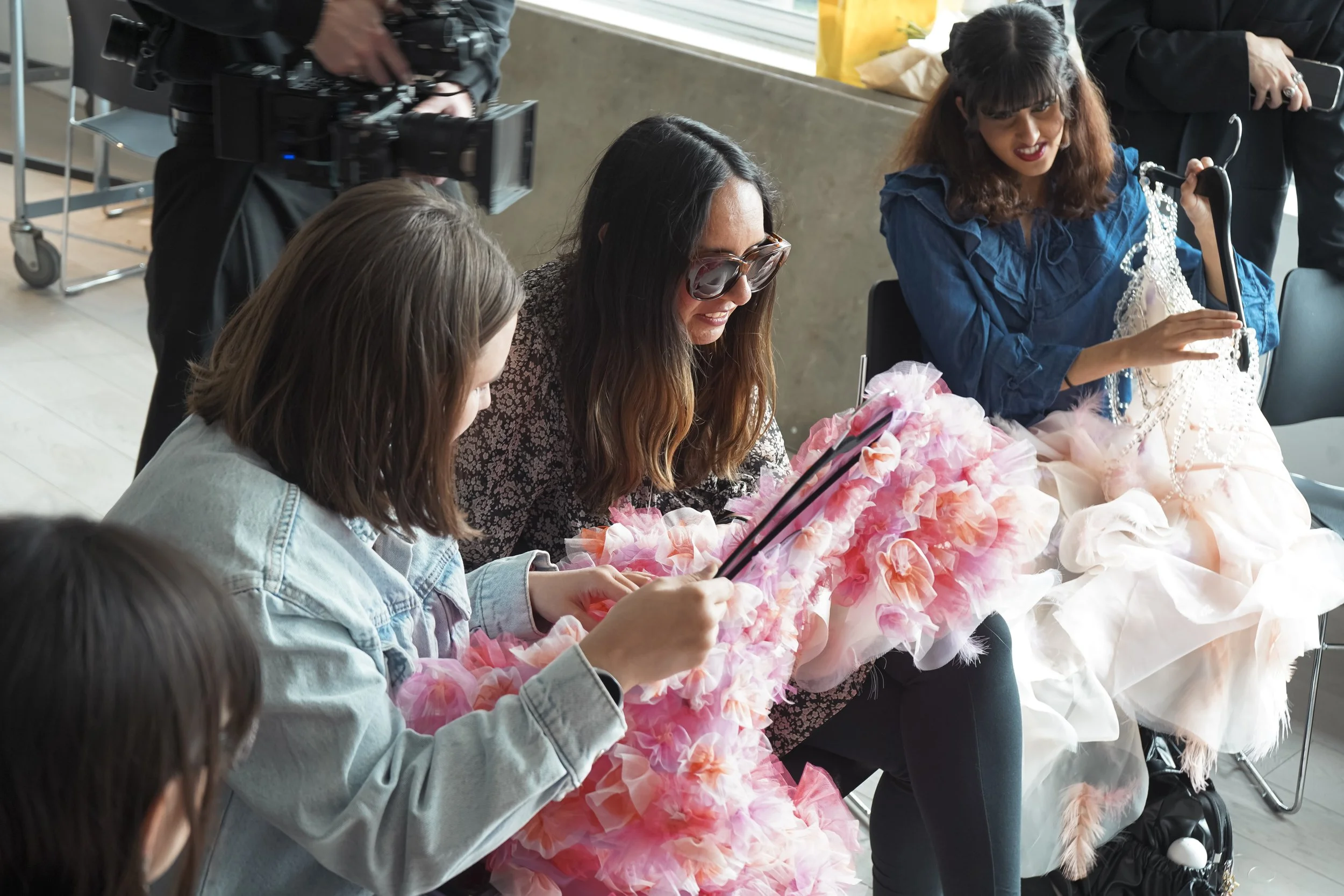 Three guests explore gowns by touch, one a pink ruffled dress and another a beaded pale pink gown, as a camera operator films.