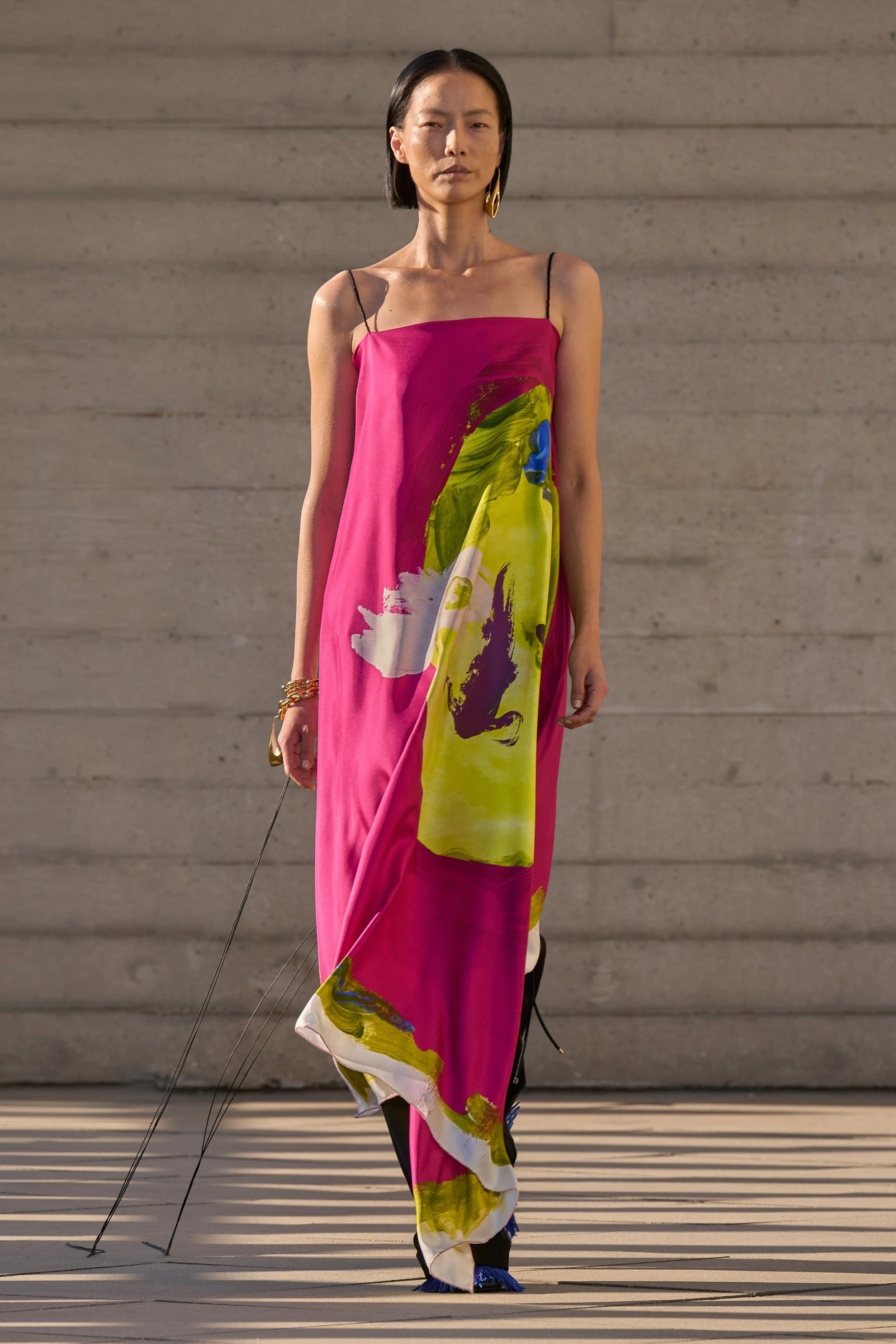 A model in a long pink slip dress with an abstract lime and purple print walks against a concrete wall.