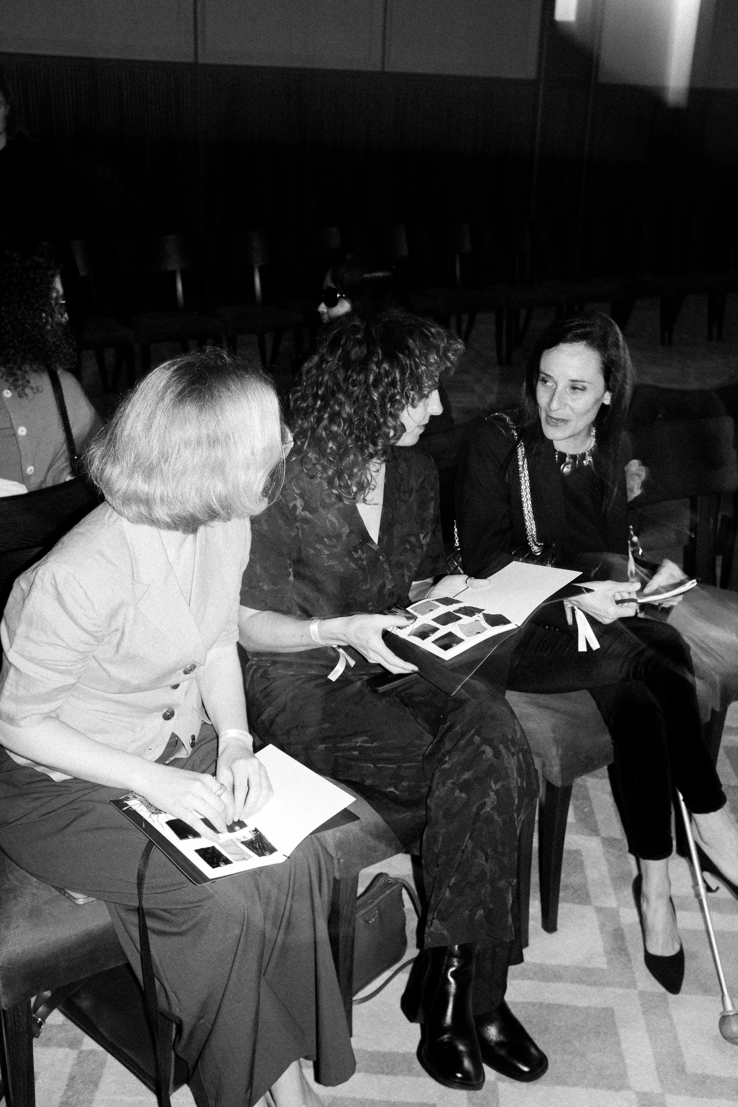 A black and white photo of three blind guests seated side-by-side at a fashion show, exchanging conversation, and dressed in smart and formal attire.