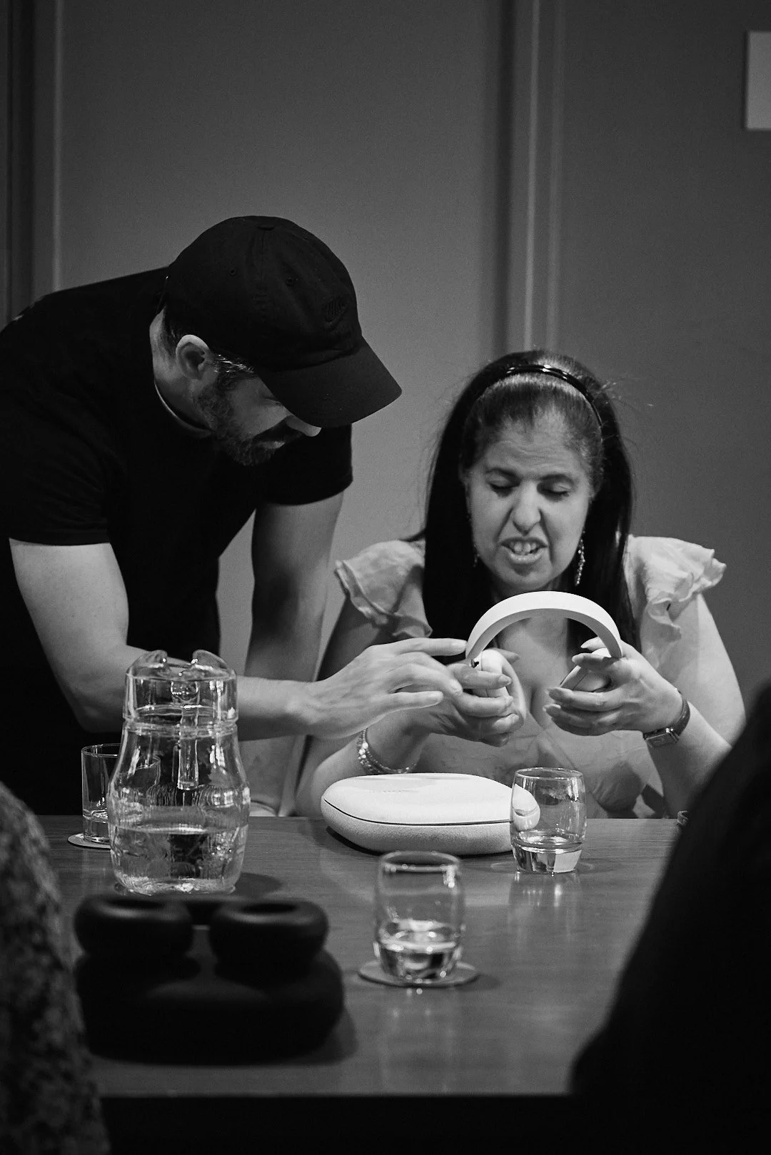 In black and white, a man in a cap guides a seated woman's hands as she explores a pair of headphones at a table.
