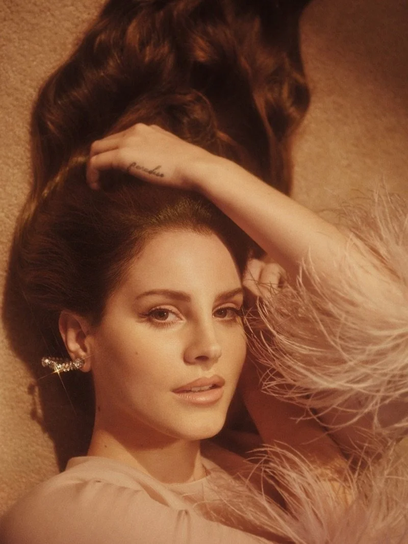An editorial photo of Lana Del Ray reclining on a beige carpet – her shiny brown hair spread out behind her. She has an elegant dress and earrings, with her hand resting on her head.