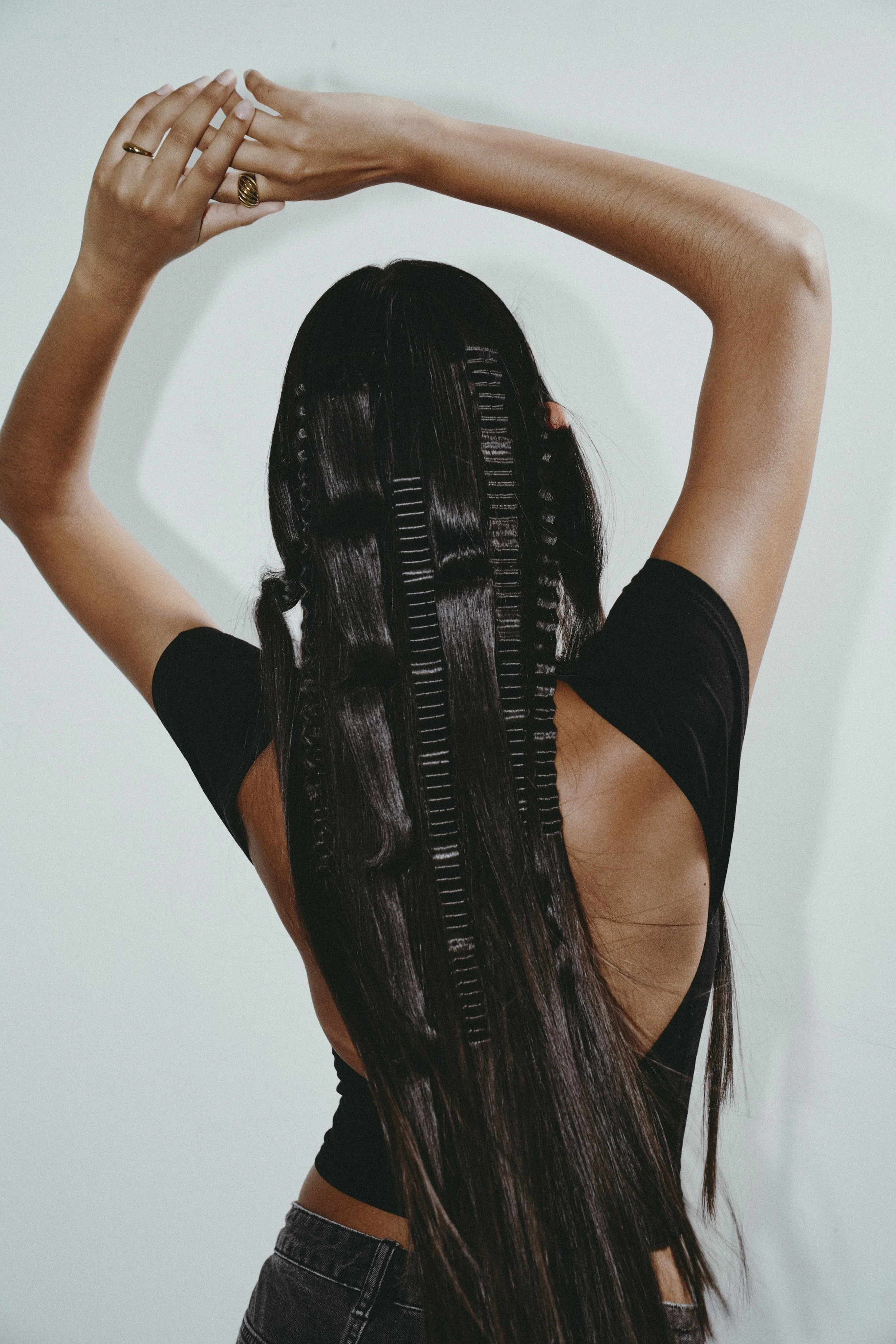 A woman with long, straight, dark hair styled with multiple small braids, with her arms raised above her head and her hands clasped, standing against a plain light background.