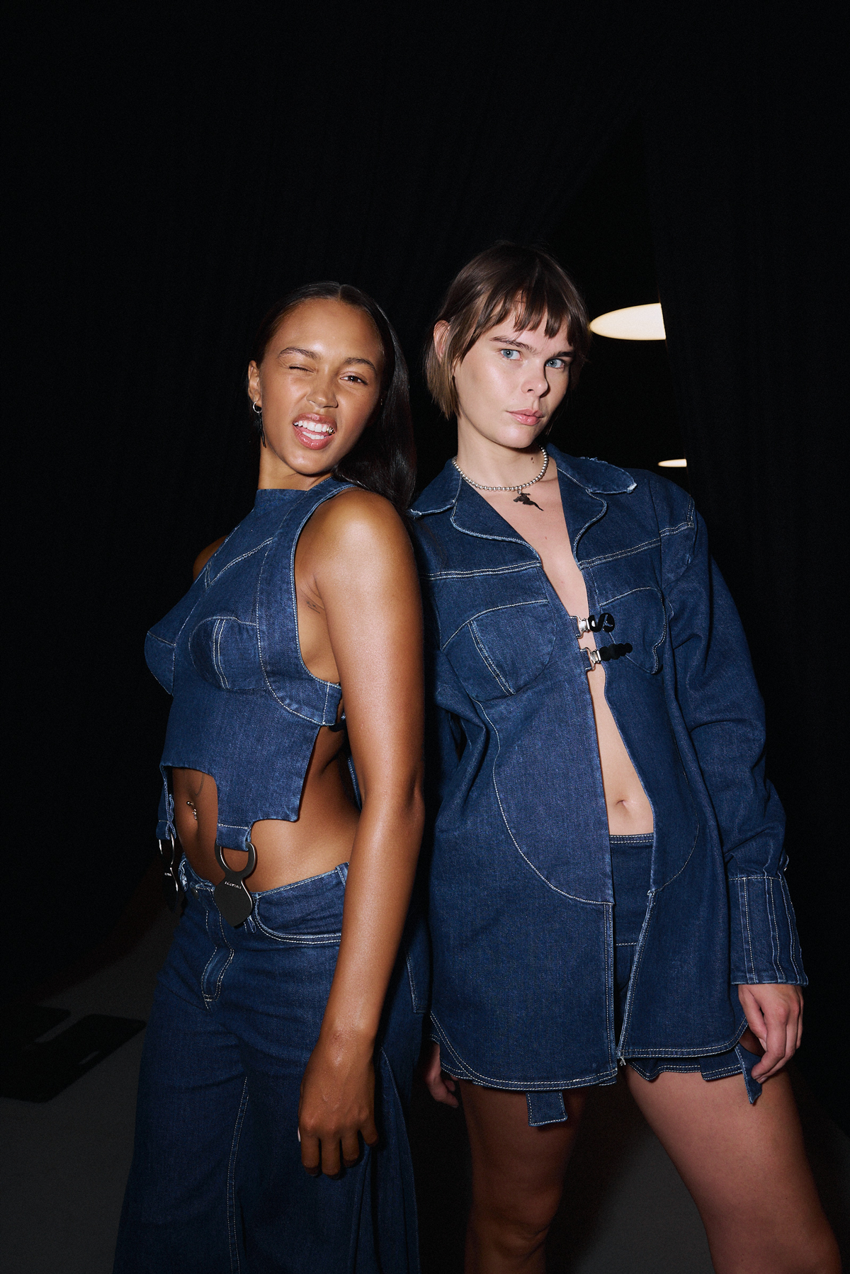 Two women wearing denim outfits posing against a dark background, both looking towards the camera