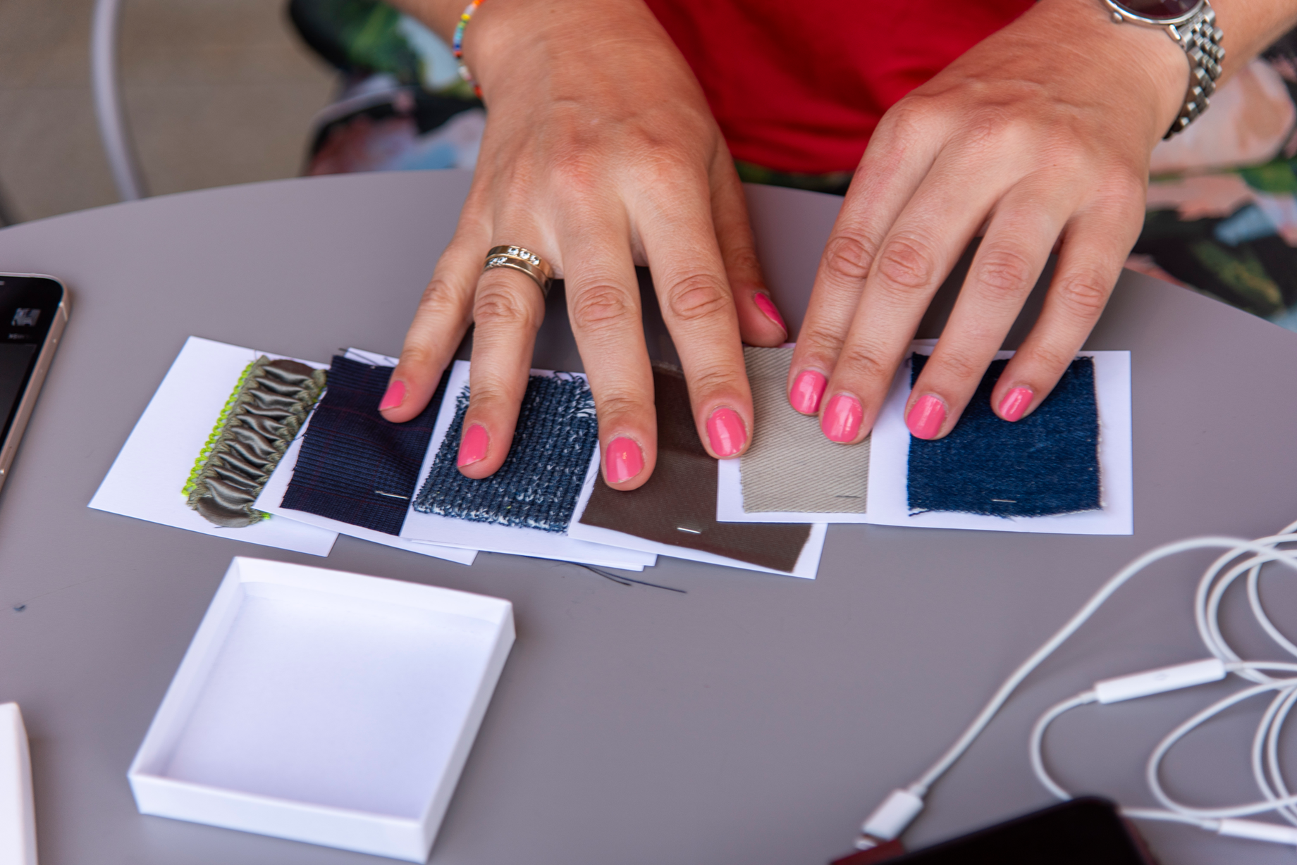 A person's hands with pink fingernail polish examining fabric swatches on paper, with a white box and electronic cords nearby.