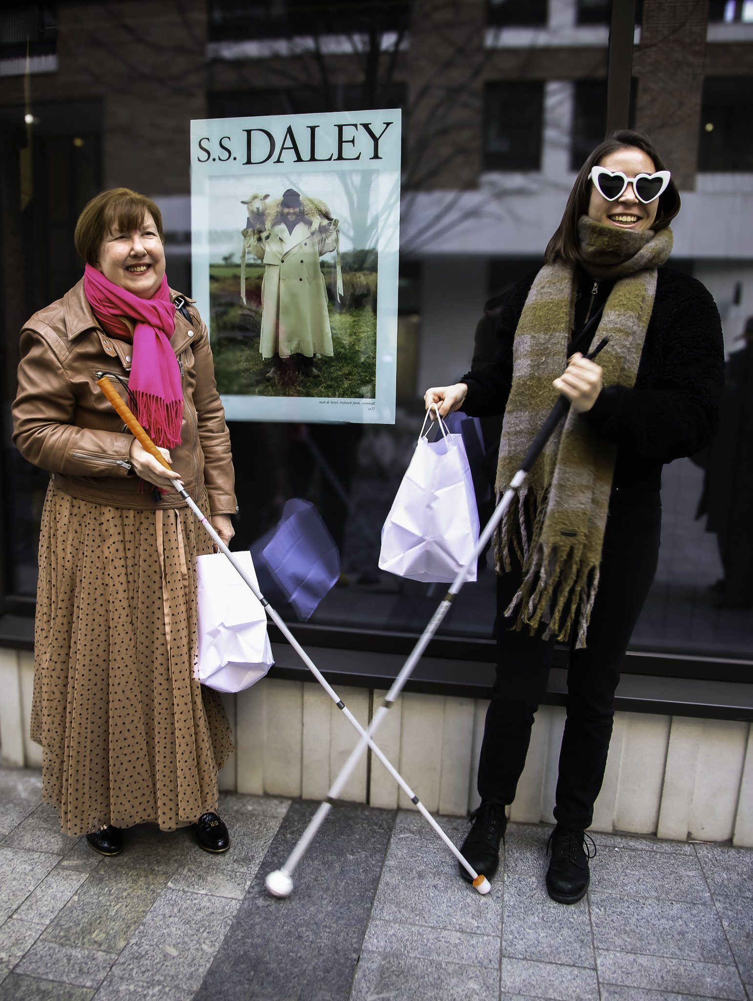 Two women standing on the pavement outside a retail window display, both holding white canes and shopping bags.