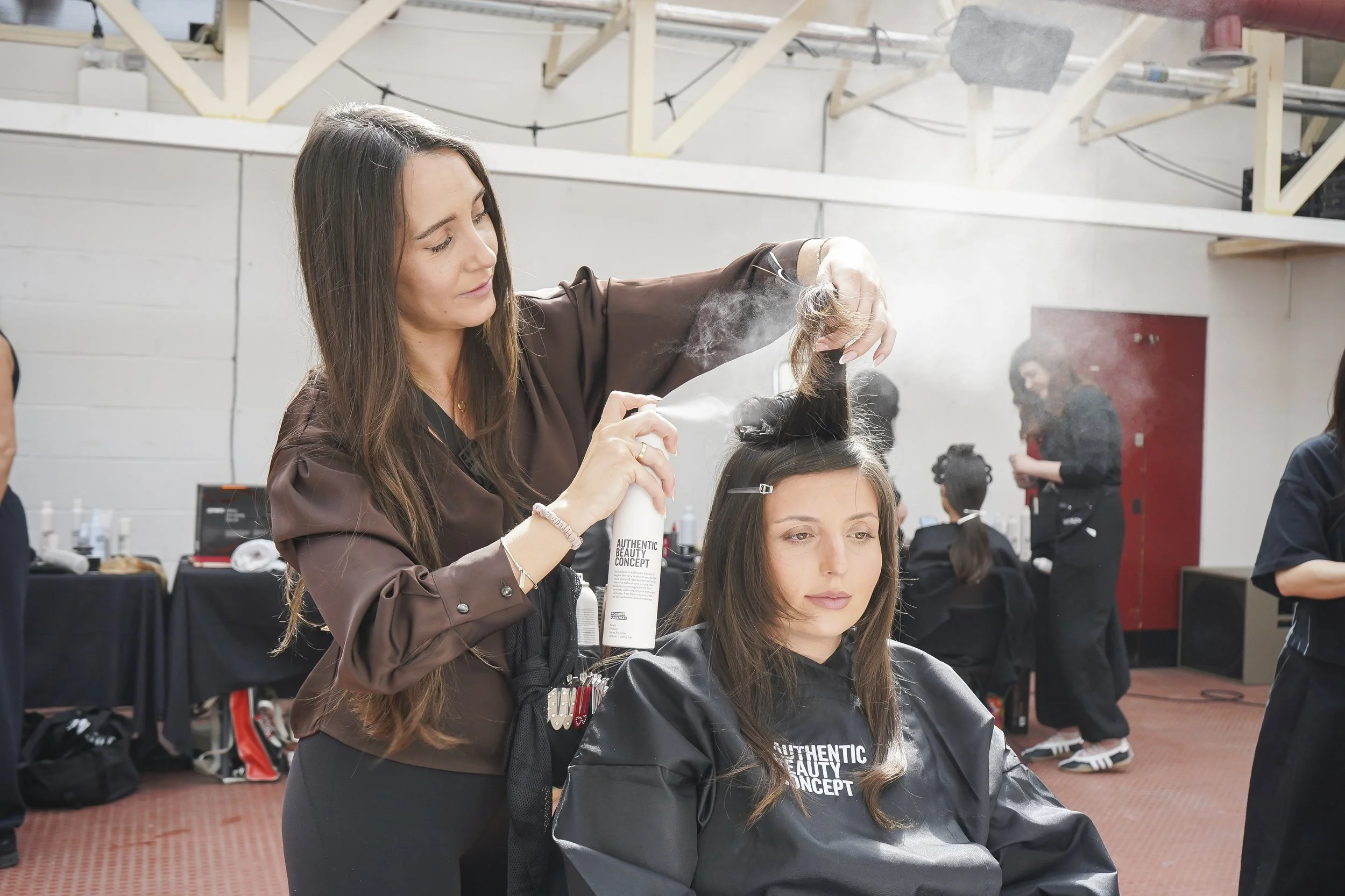 A hairstylist applies hair spray to a woman's hair in a salon setting, with other people and salon equipment visible in the background.