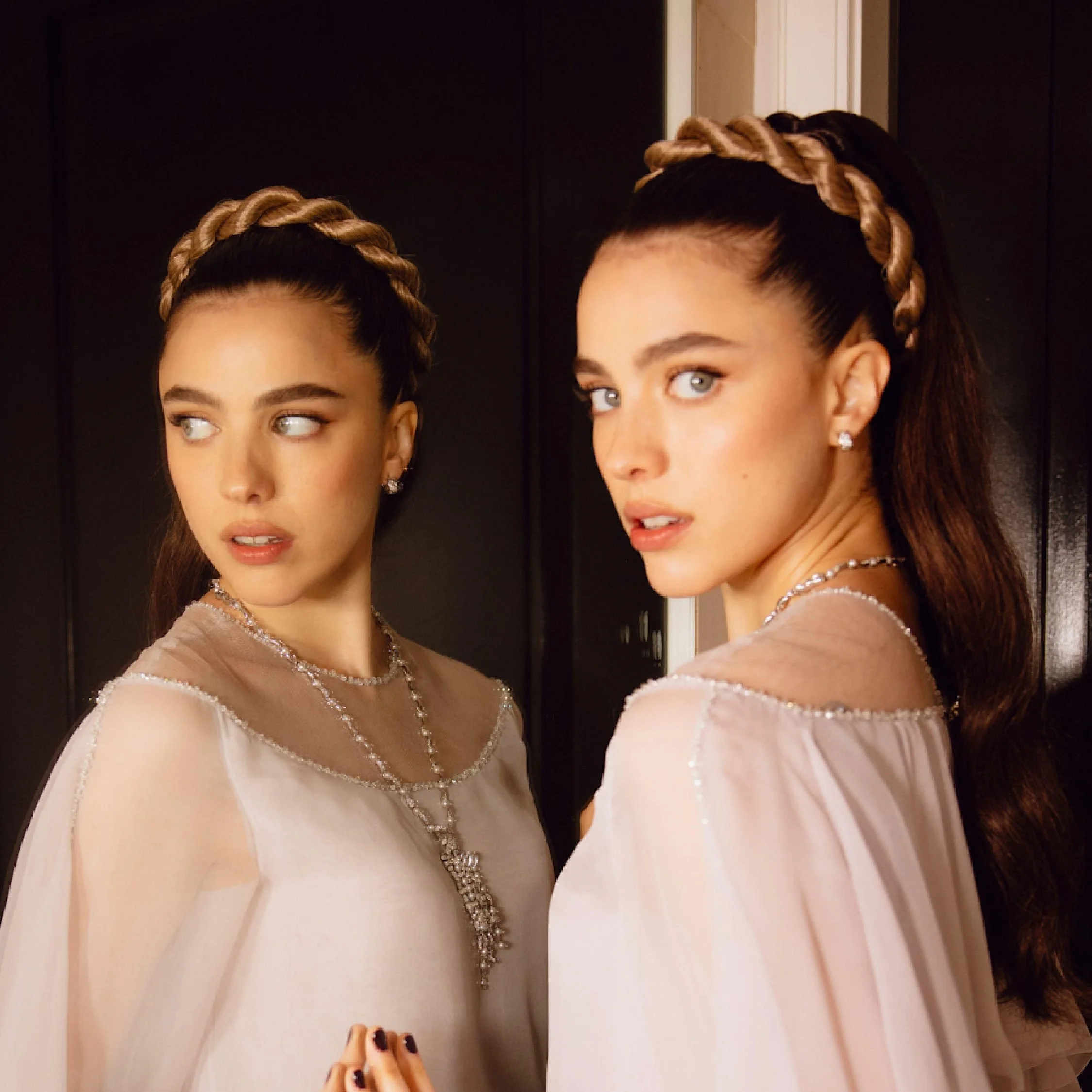 Actress Margaret Qualley has her hair styled in an elegant up-do with a braided crown, looking away, wearing diamond earrings, a beige dress with beaded details, and a pearl necklace, standing in front of a mirror.