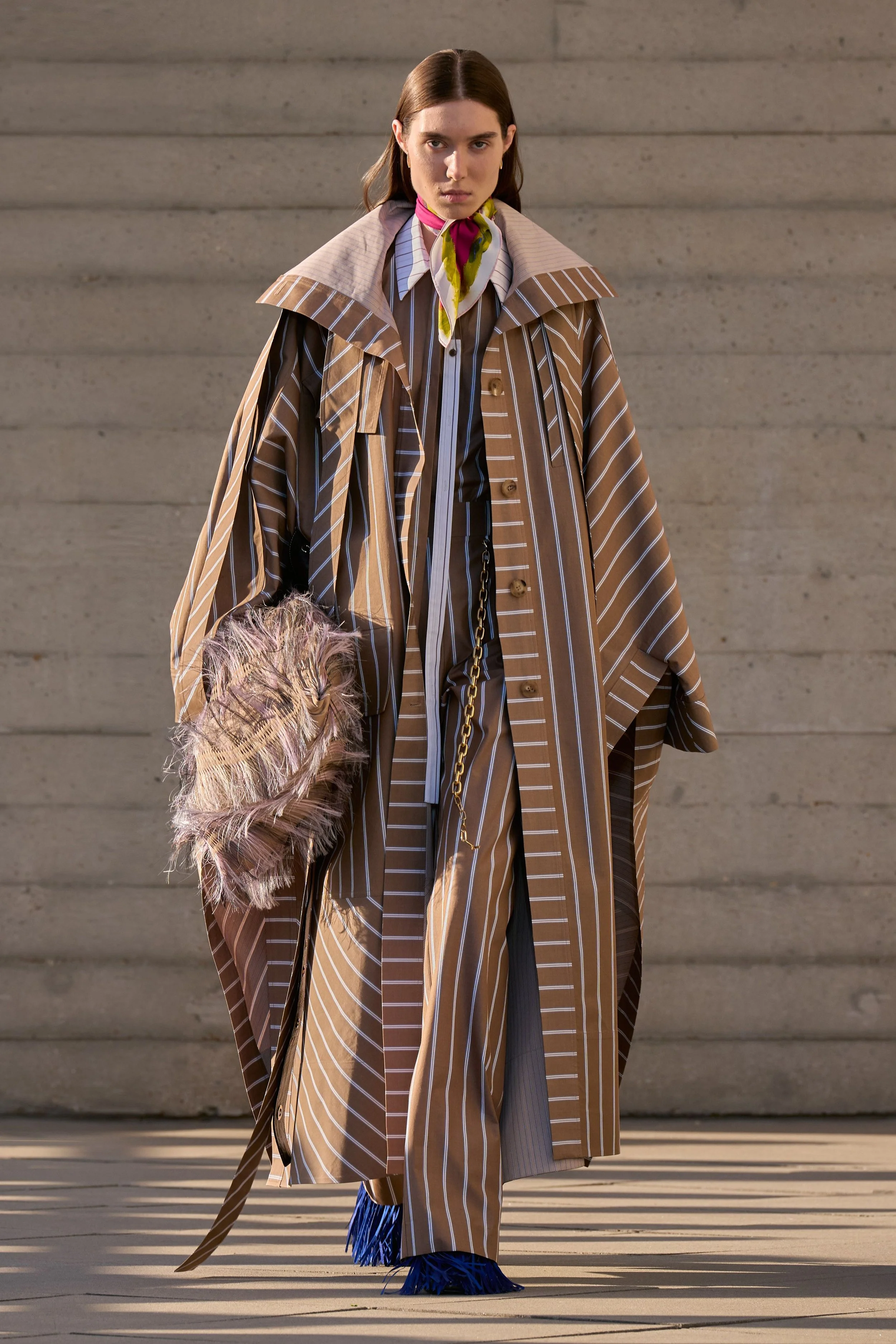 A model in an oversized brown-and-white striped coat and trousers carries a fringed bag.