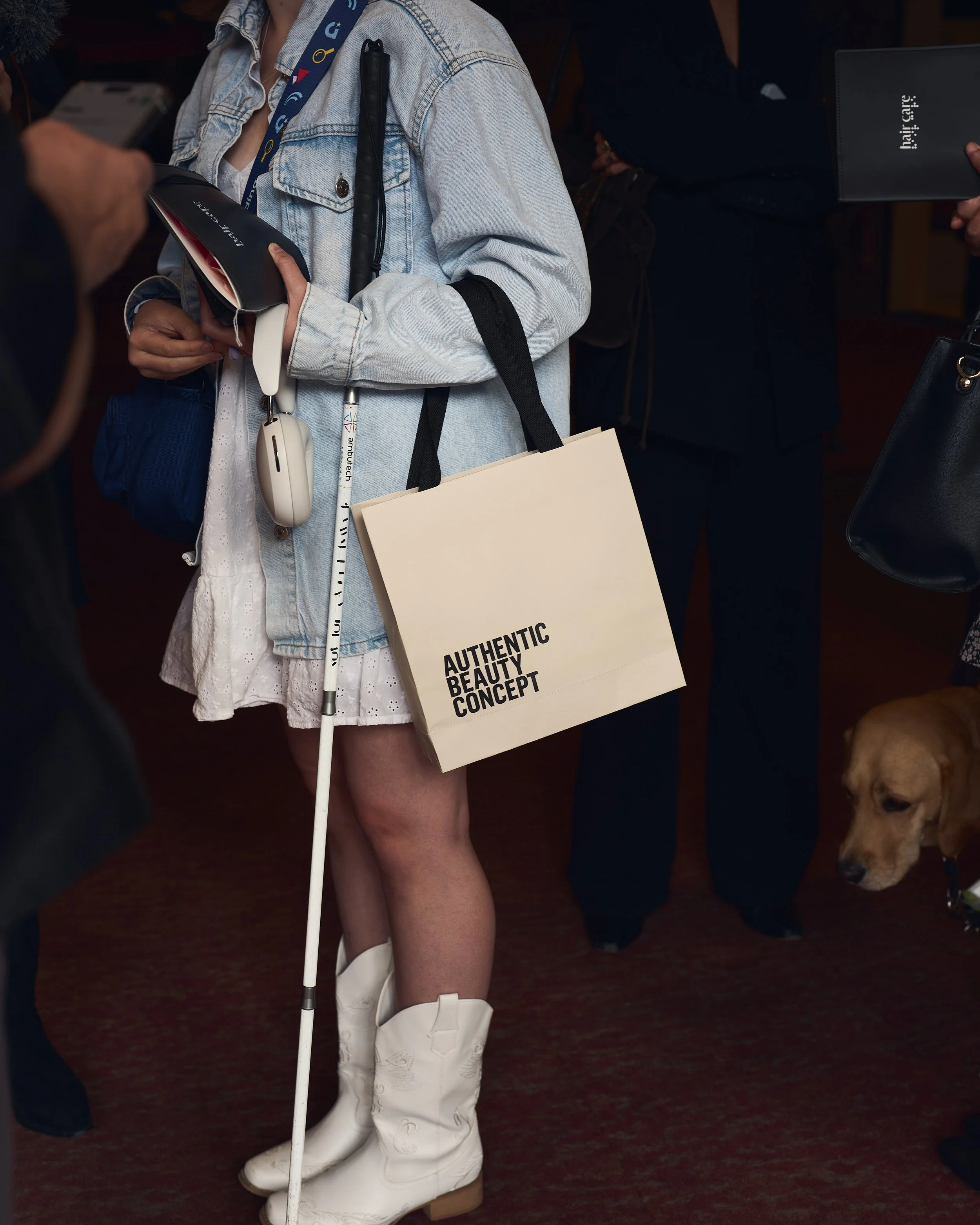A guest with a white cane, white cowboy boots and an Authentic Beauty Concept bag holds a Hair & Care booklet.