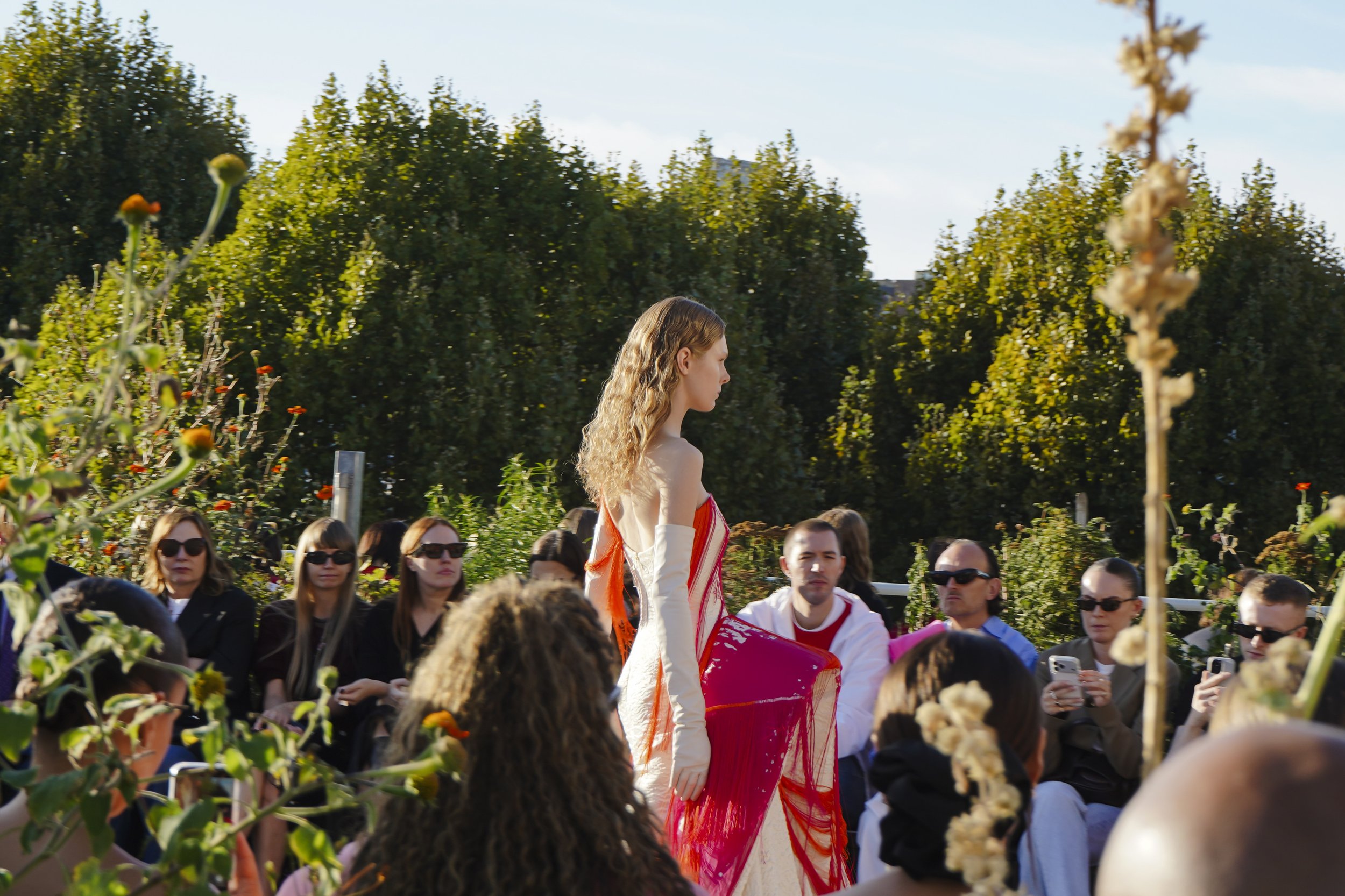A model in a strapless ivory gown with red and pink fringe walks past guests in a sunlit rooftop garden.
