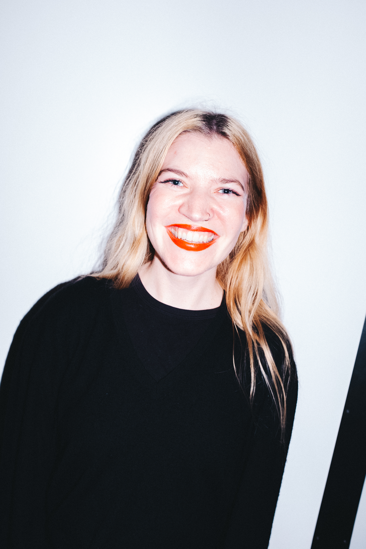 A young woman with long blonde hair, wearing a black top, smiling with bright red lipstick, and standing against a plain white background.