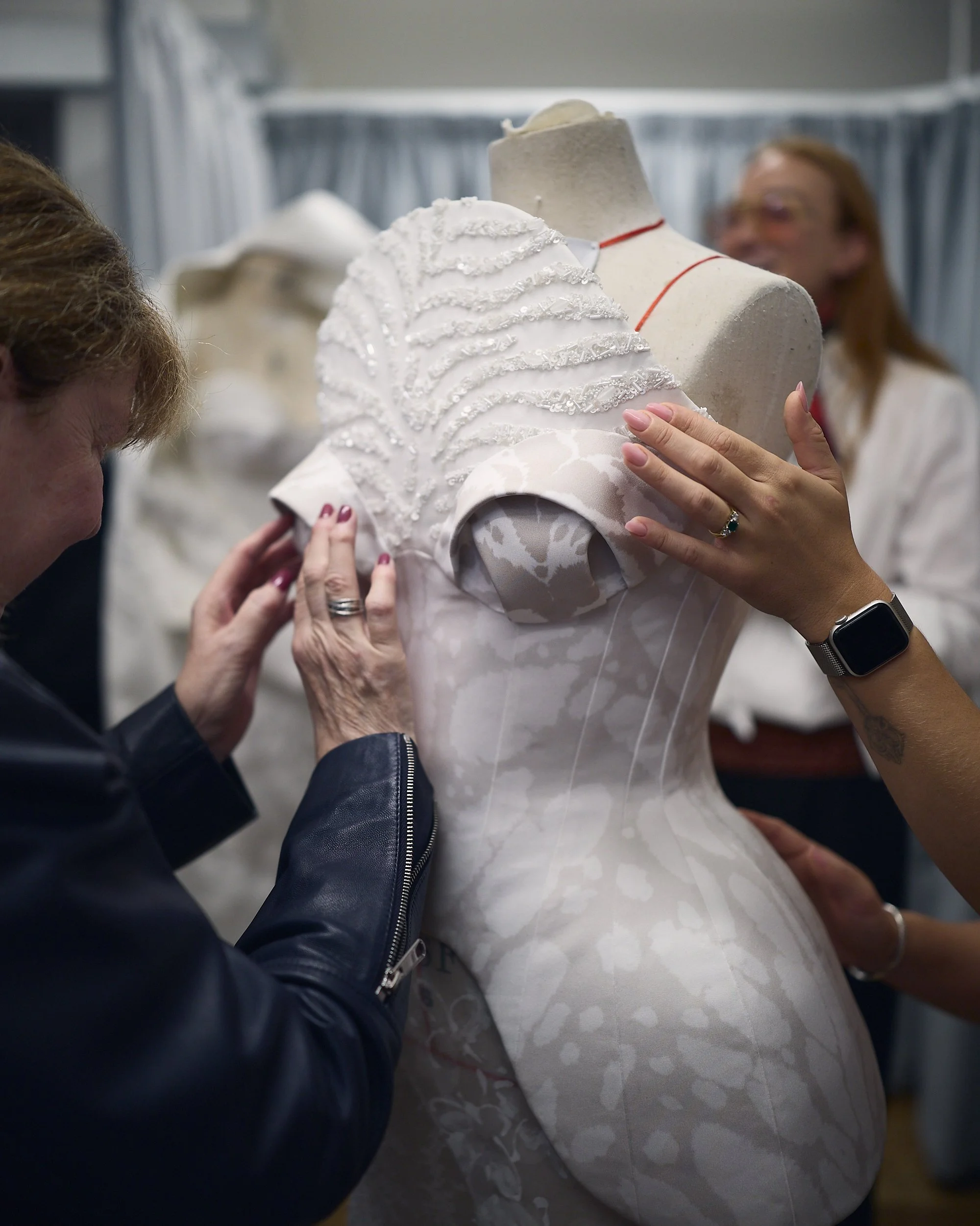 Two guests explore a sculpted white corset gown with a beaded shoulder by touch, as it stands on a mannequin.