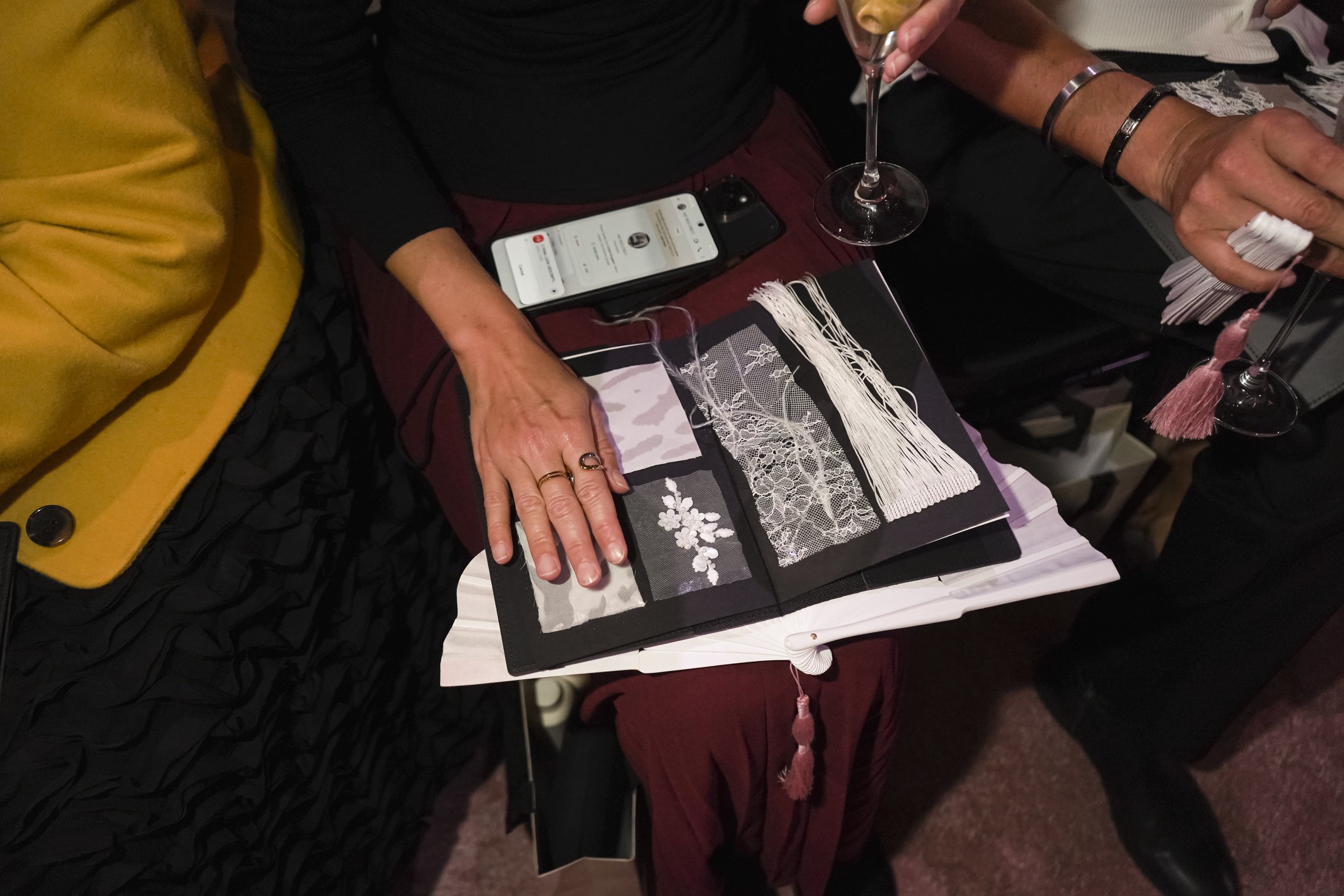 A guest's hand rests on a tactile booklet of lace, beaded floral and fringe swatches, with a white fan on her lap.