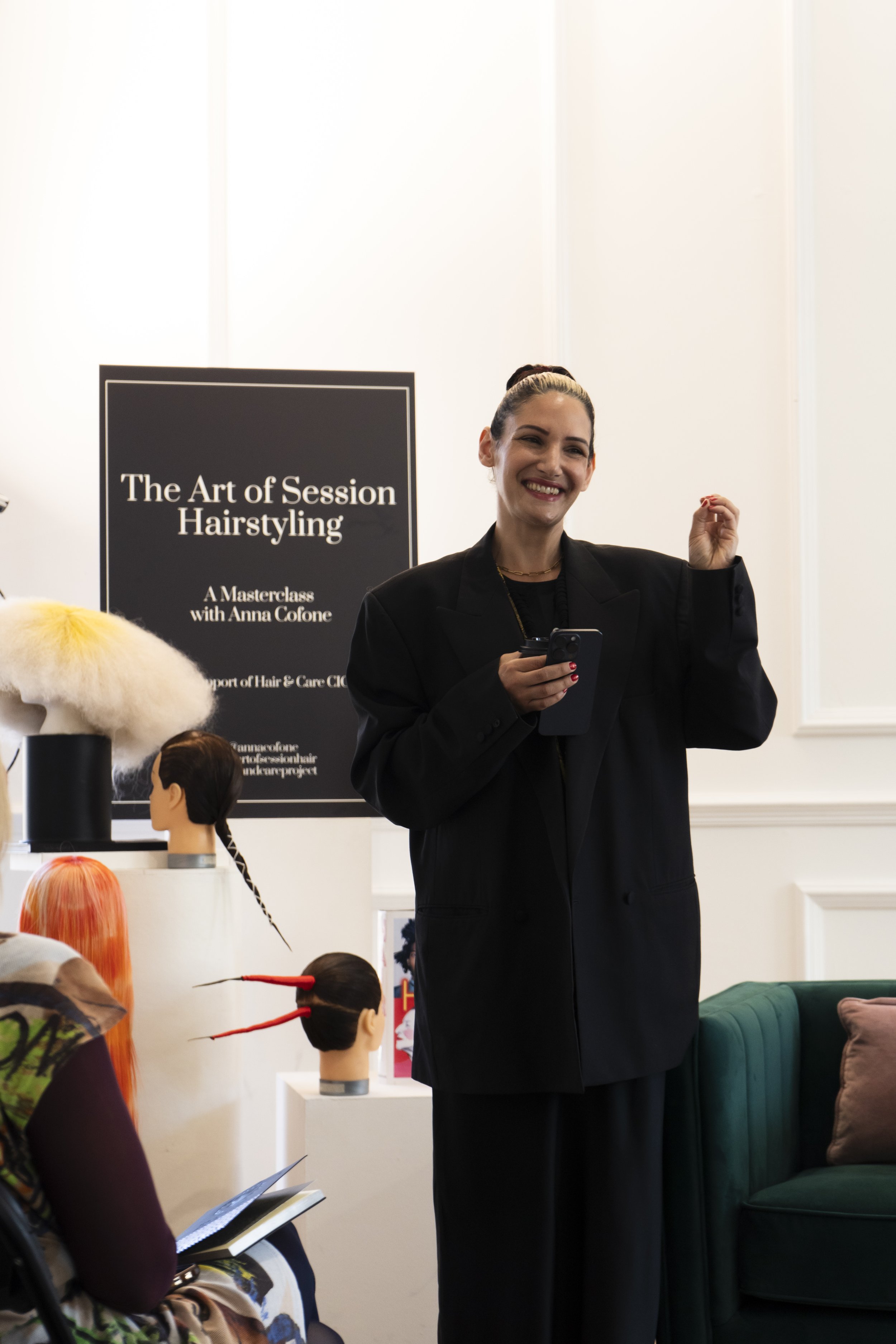 A woman in black clothing smiling, standing near a display with mannequins and wigs.