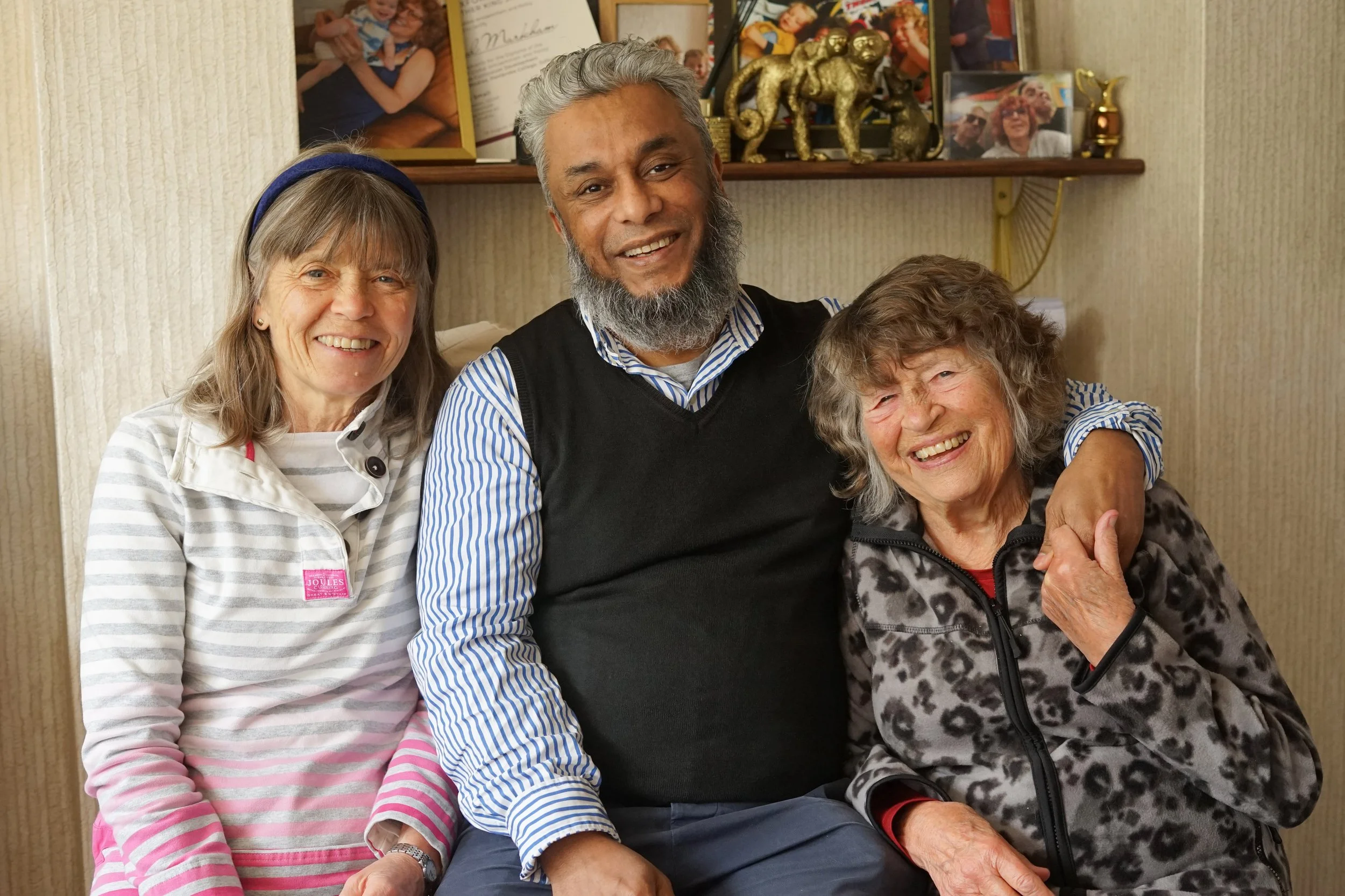 A group of four people, three women and one man, smiling and sitting close together in a cozy indoor setting. They appear to be happy and enjoying each other's company.