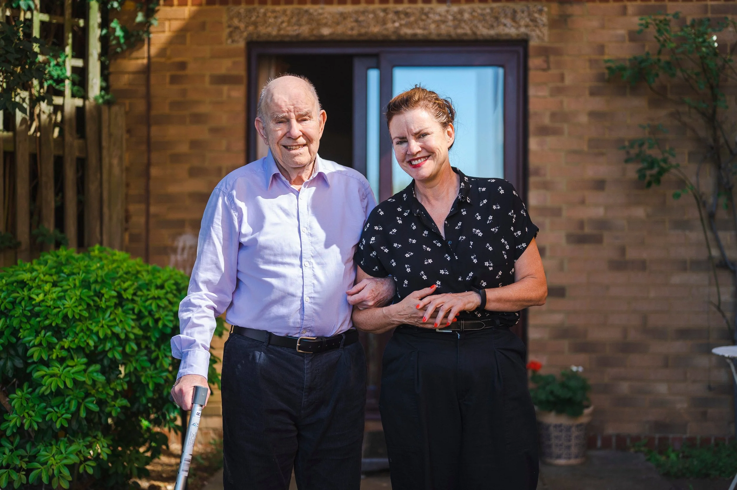 An elderly man using a cane and a woman with red hair smiling, standing outside in front of a brick house with green bushes and trees.