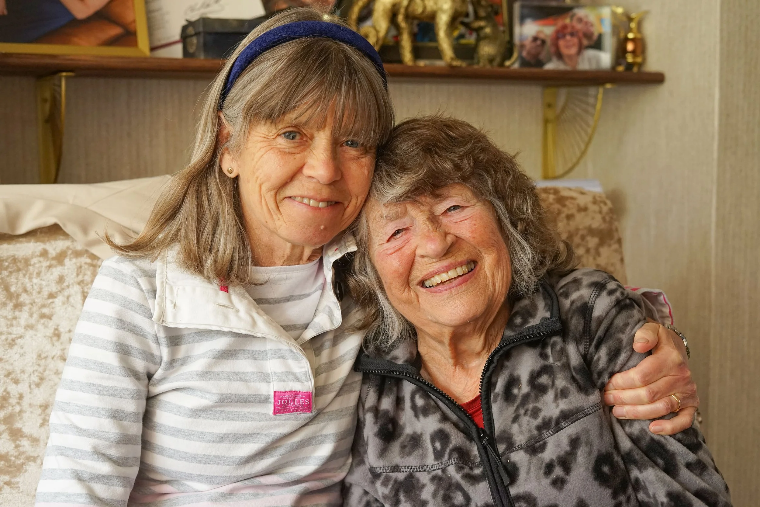 Two older women hugging and smiling, sitting on a couch in a cozy living room. One woman has light hair with a blue headband, and the other has curly gray hair and is wearing a black zip-up jacket. In the background, there is a shelf with decorative items and a framed photo of two women.