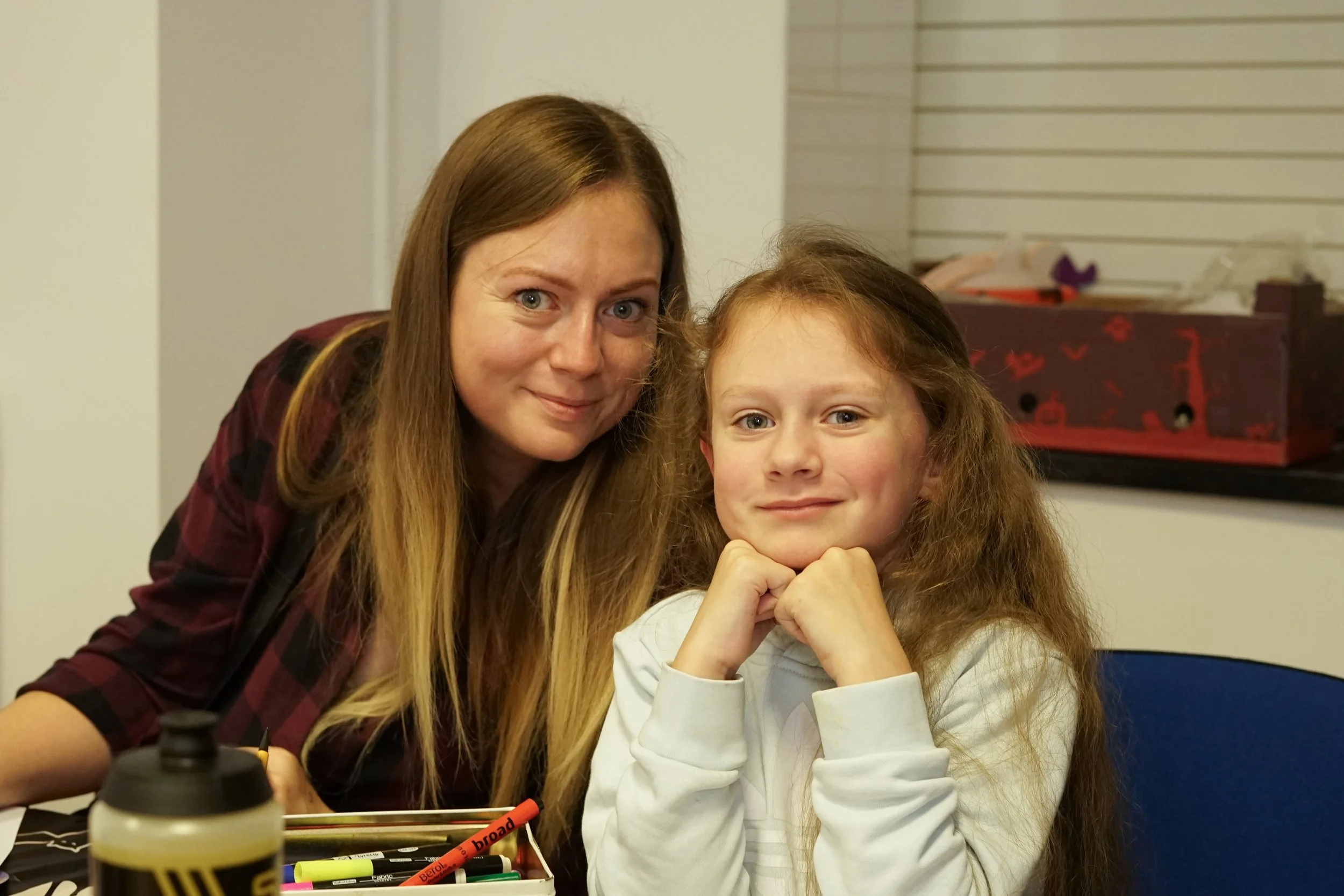 A woman with long brown hair and a young girl with curly red hair sit close together at a table, smiling at the camera. The table has colorful markers, a glue bottle, and a box of drawing supplies.