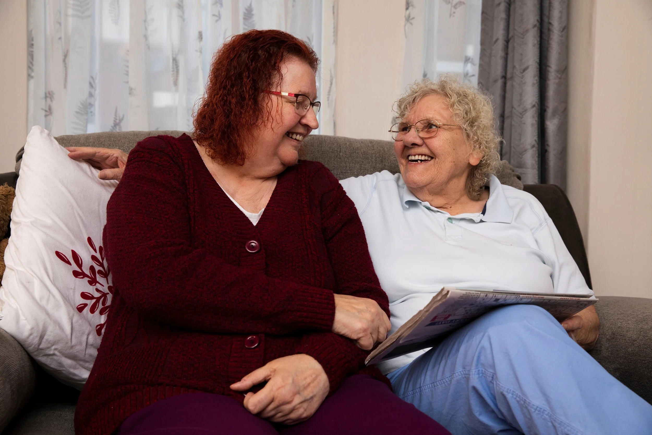 Two women, one elderly with gray curly hair and glasses, and one middle-aged with red curly hair and glasses, sitting on a sofa, smiling and looking at each other, with a window and curtains in the background.