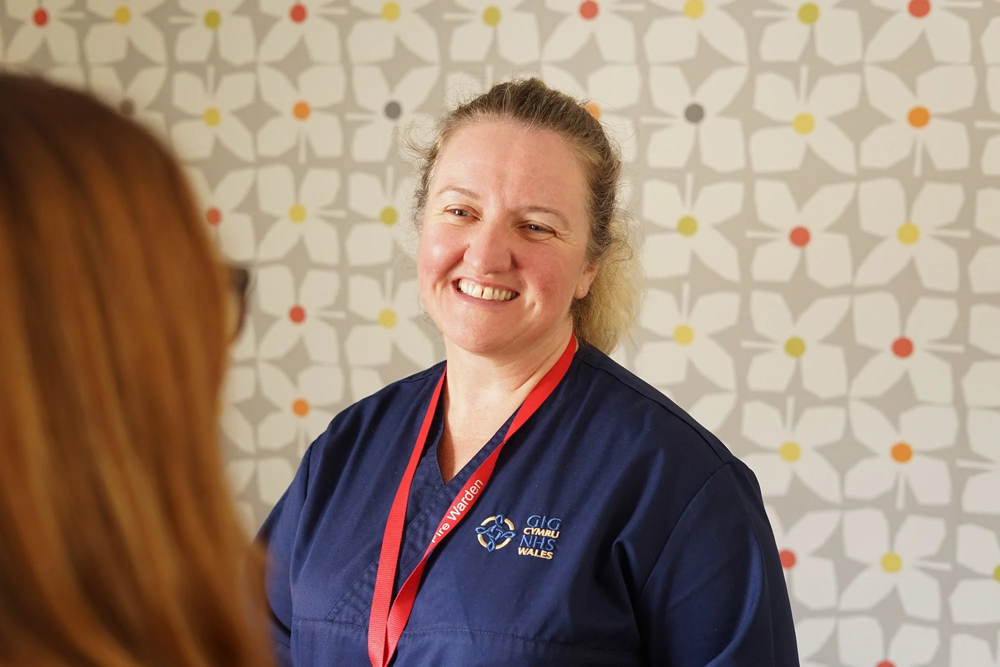 A woman with blonde hair smiling, wearing a dark blue uniform with the logo of GIG Cymru Wales, standing in front of a patterned background, with a person with red hair partially visible in the foreground.