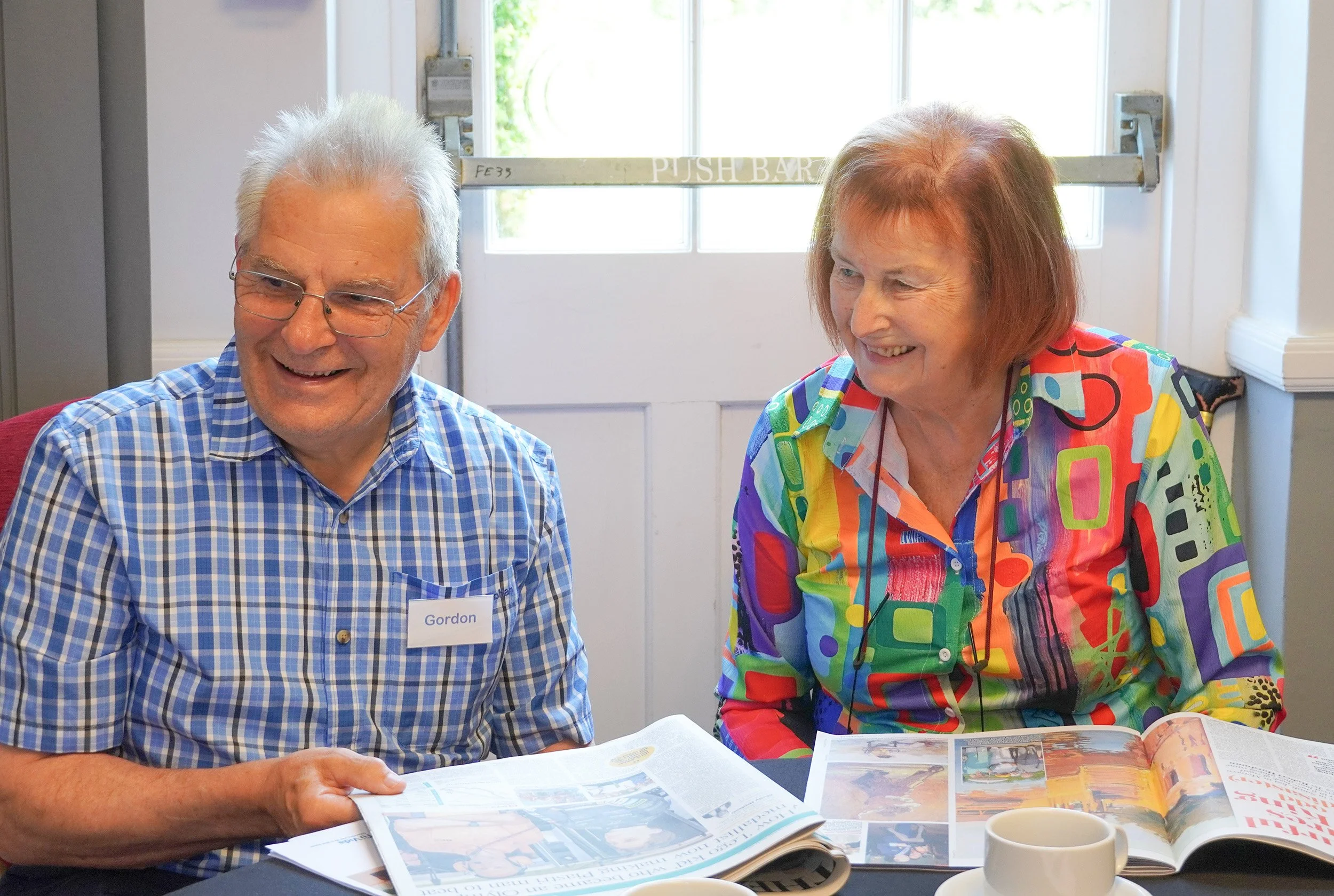 An elderly man with white hair, glasses, wearing a blue checkered shirt and a name tag that reads 'Gordon,' sitting at a table with an open magazine. Beside him is an elderly woman with short reddish hair, wearing a colorful, geometric-patterned shirt, smiling while reading a magazine. They are in a bright room with a window in the background.