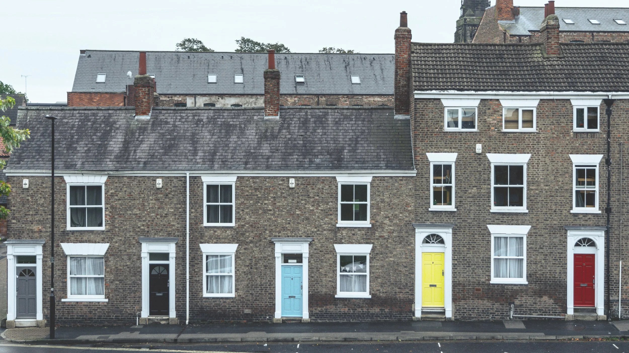 Row of brick townhouses with colorful front doors and white-framed windows, street with parking spaces and a black street lamp, some buildings with visible chimneys and sloped roofs.