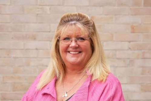A smiling woman with blonde hair and glasses, wearing a pink cardigan, standing in front of a brick wall.
