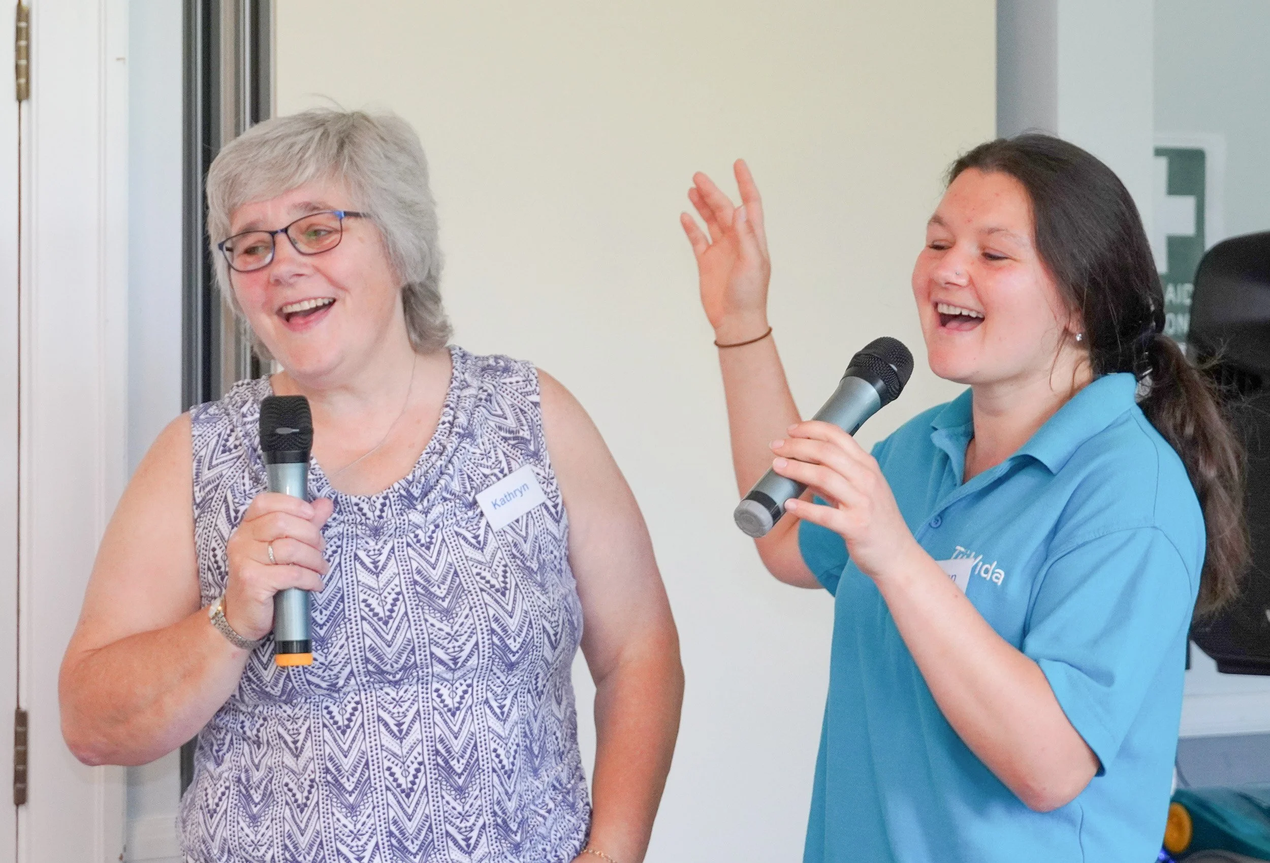 Two women smiling and talking into microphones during an event. The woman on the left has gray hair, glasses, and is wearing a patterned sleeveless top, while the woman on the right has dark hair tied back and is wearing a blue polo shirt.