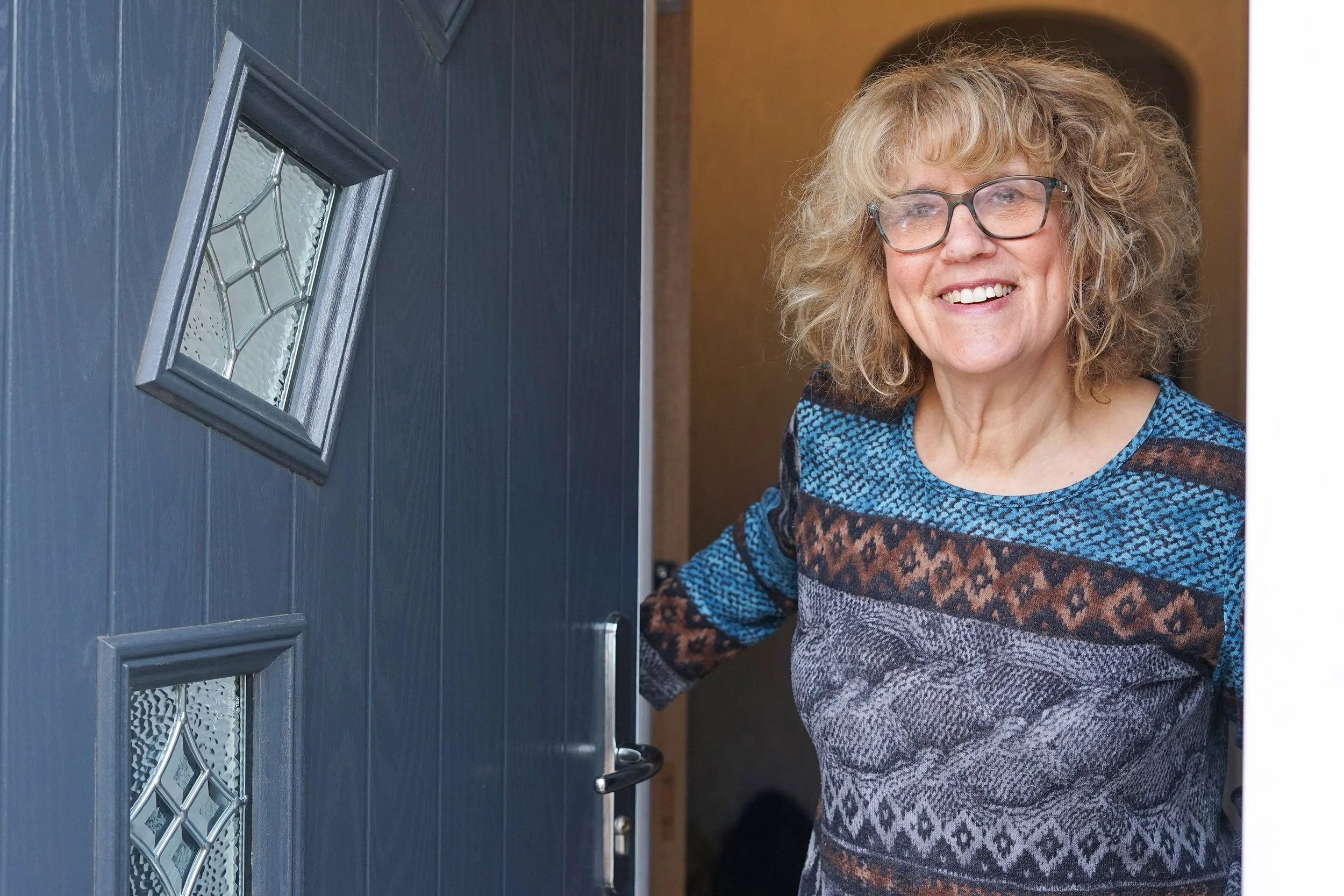 An elderly woman with curly blond hair and glasses smiling as she stands at her front door, about to enter her house.