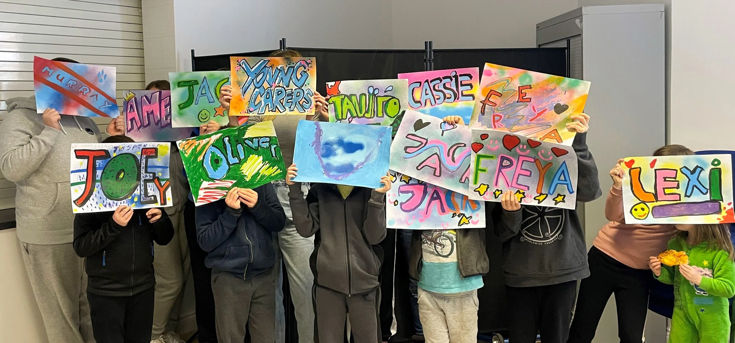 Children holding colorful handmade signs with names and drawings at an indoor event.