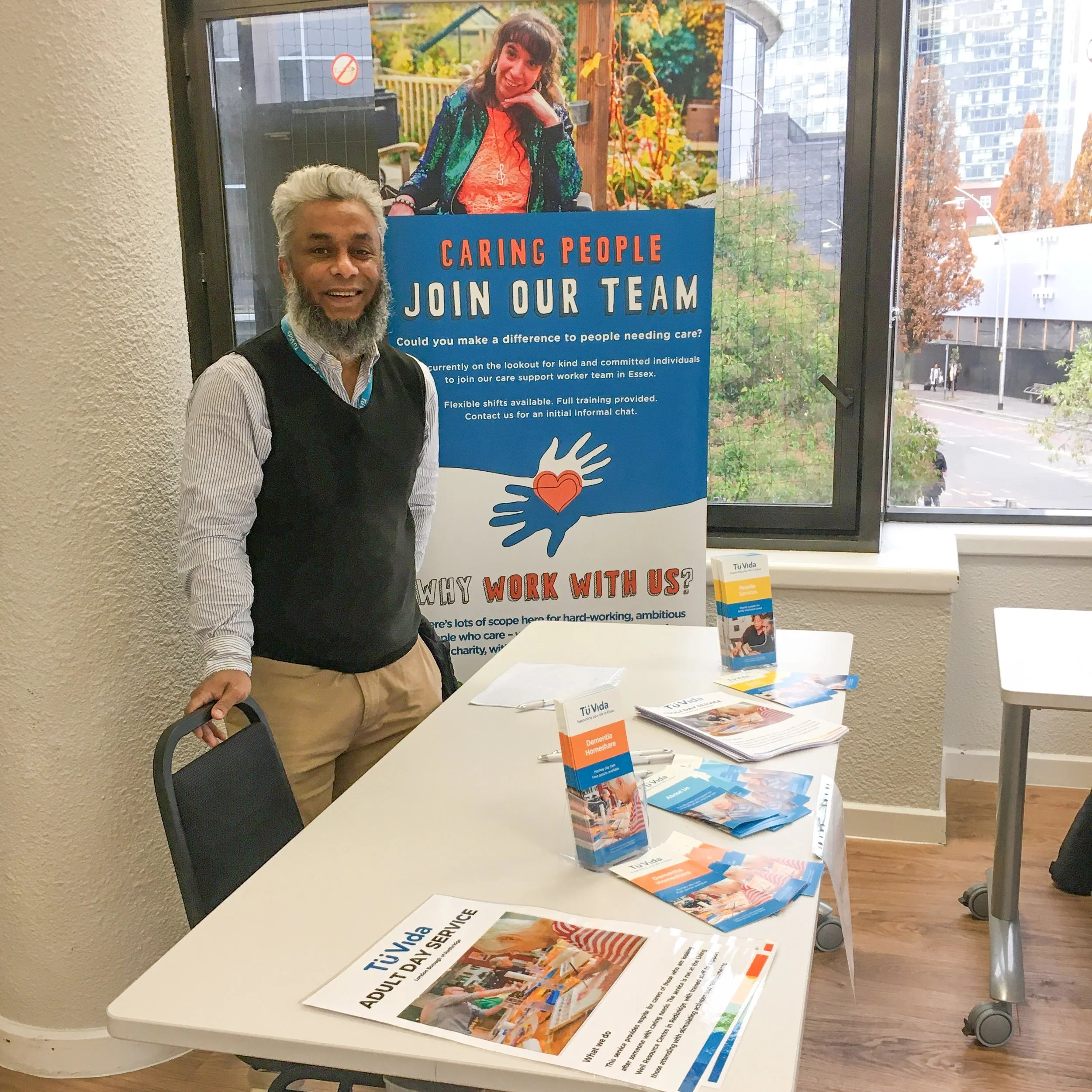 A man with white hair and a beard, wearing a striped shirt and black vest, standing at a table with brochures. Behind him is a banner with a photo of a woman with long curly hair, smiling, and the text 'CARING PEOPLE JOIN OUR TEAM'. The table and brochures are related to the organization's caregiver services.