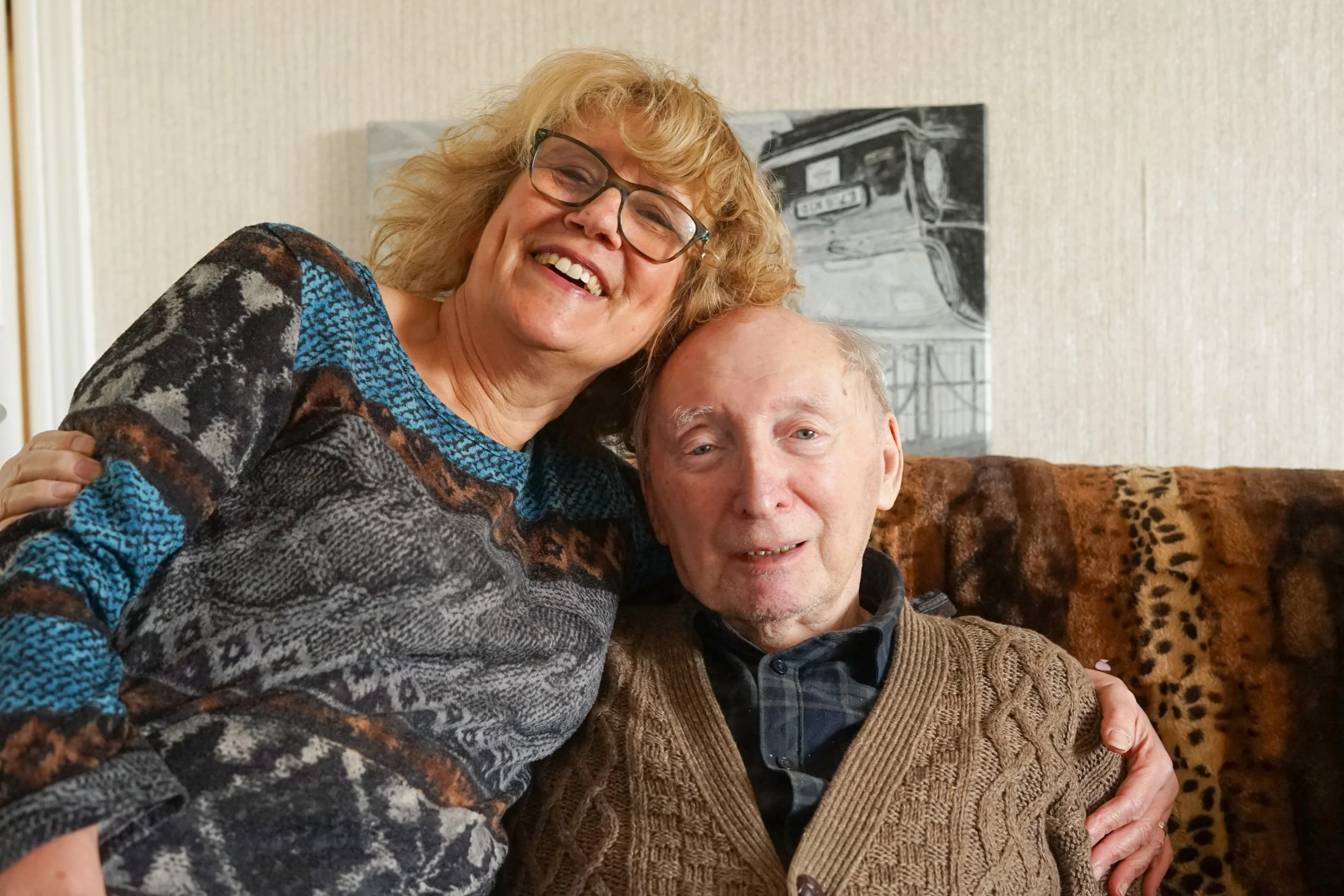 A cheerful woman with curly blonde hair, wearing glasses and a colorful patterned sweater, hugging an older man with gray hair, wearing a brown knitted cardigan and a dark shirt. They are sitting on a leopard print couch in a cozy room with a textured light-colored wall and a black-and-white painting of a vintage car behind them.