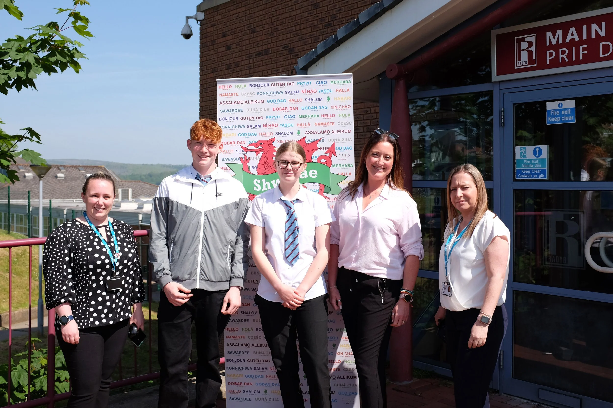 Group of five people standing outdoors in front of a sign with multilingual greetings and a dragon illustration, near a building with a blue door and a fire exit sign.