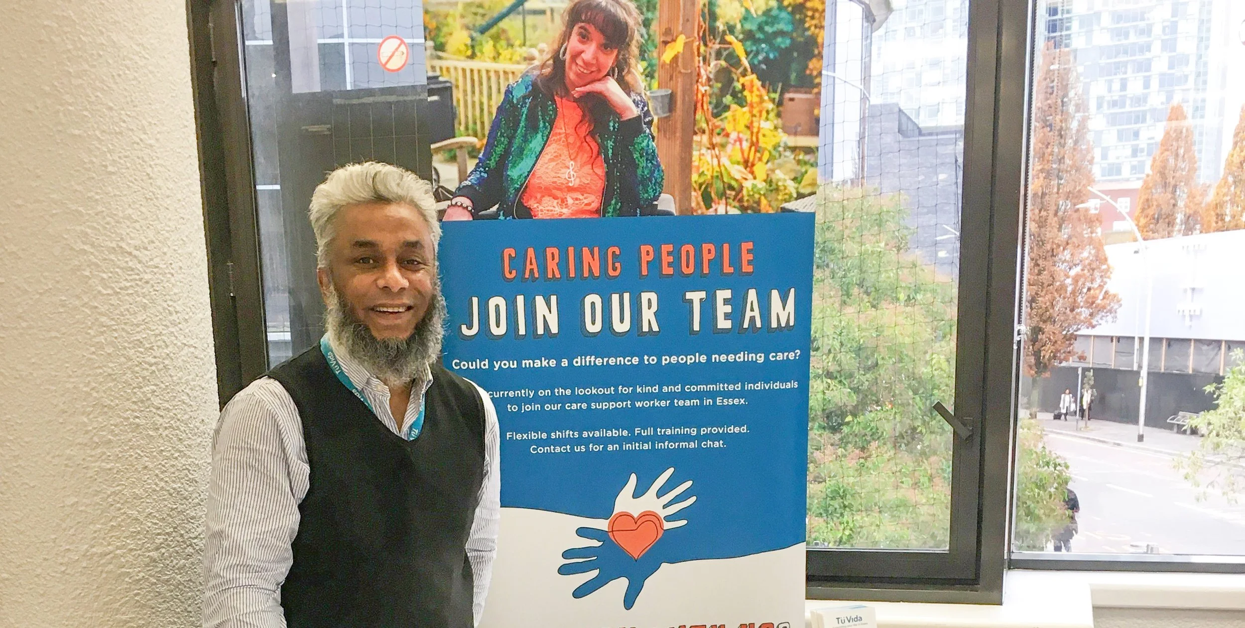 A man with gray hair and a beard standing next to a care support recruitment poster. The poster features a woman smiling outdoors, with trees and a wooden fence in the background, and text inviting people to join the care team.