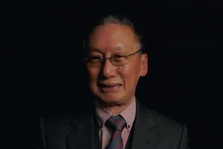 Portrait of an elderly man in a suit and tie with glasses, smiling against a black background.