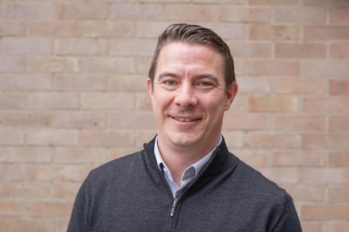 A man smiling, wearing a dark zip-up sweater and a collared shirt, standing in front of a brick wall.
