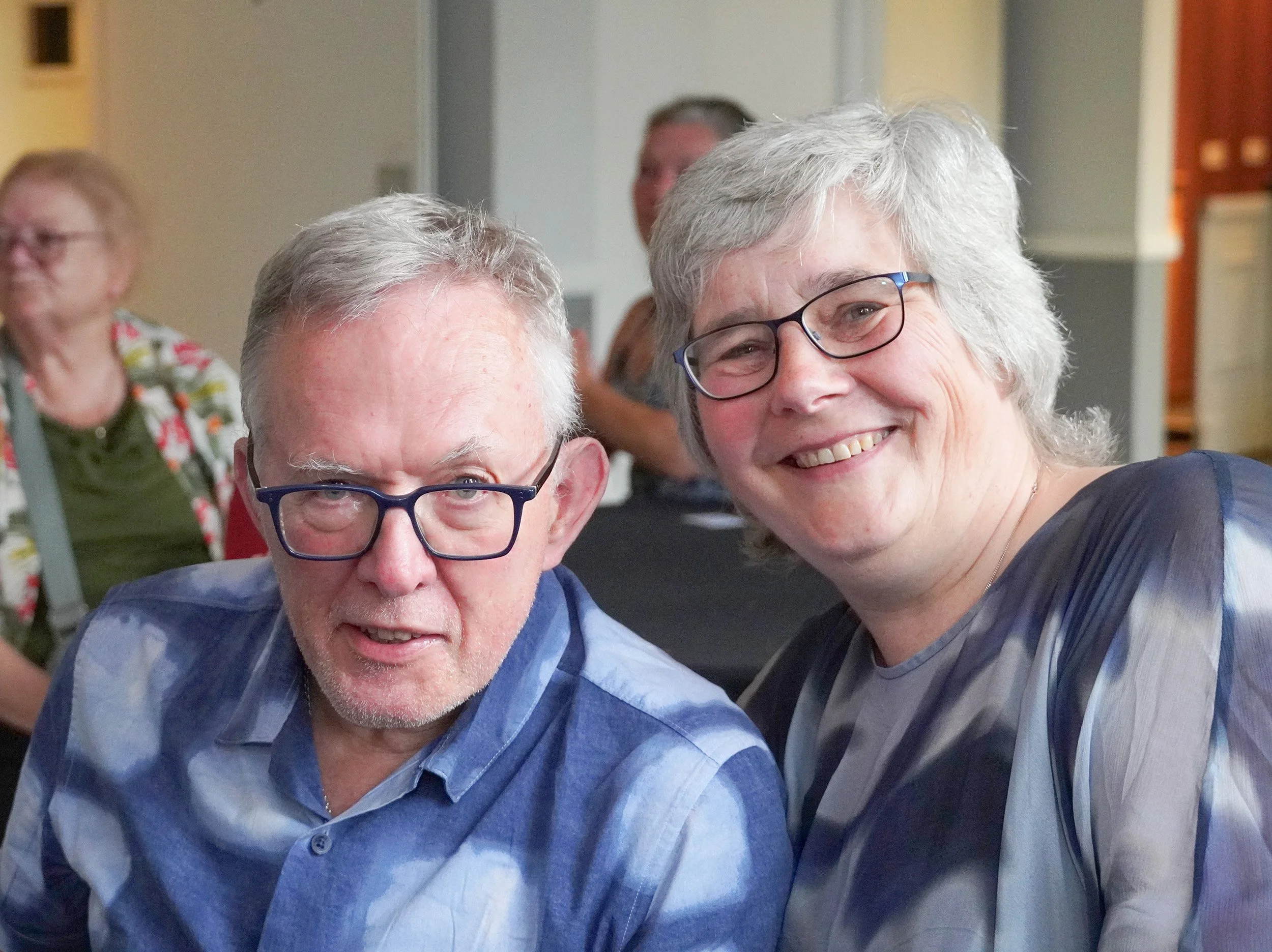 Smiling elderly woman with gray hair and glasses, sitting with an elderly man with gray hair and glasses in a social setting. In the background, other seniors are visible, some wearing colorful attire. The overall scene depicts a happy gathering or event.