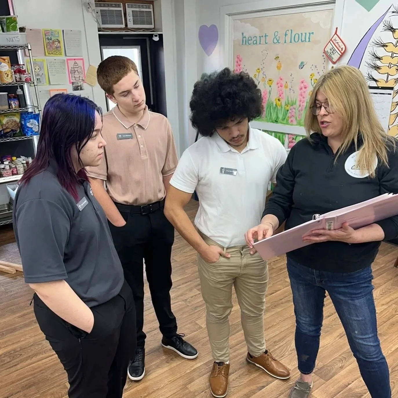 Three young adults and an adult woman are gathered around, looking at a binder together inside a room with wooden flooring and colorful wall art.