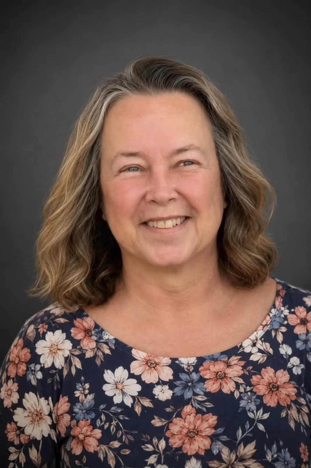 A middle-aged woman with shoulder-length wavy hair and light skin smiling at the camera, wearing a navy blue floral blouse against a dark gray background.