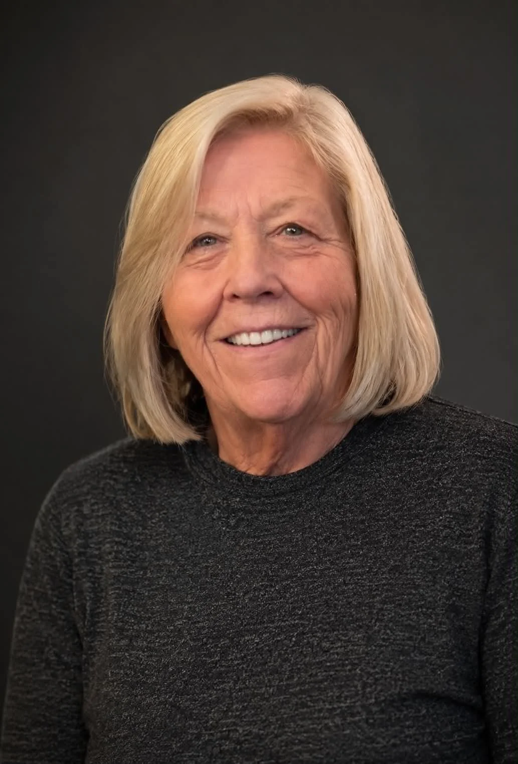 A smiling elderly woman with shoulder-length blonde hair, wearing a dark gray textured sweater, posing against a black background.