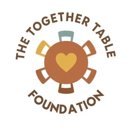Logo of the Together Table Foundation featuring a central orange circle with a yellow heart, surrounded by five multicolored chairs, and the text 'The Together Table Foundation' curved around the logo.