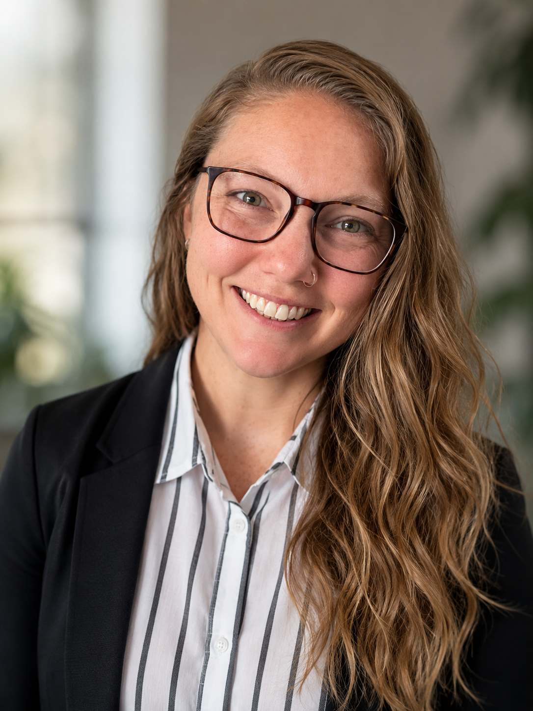 A woman with long, wavy, light brown hair, wearing glasses, a black blazer, and a white striped shirt, smiling in an indoor setting.