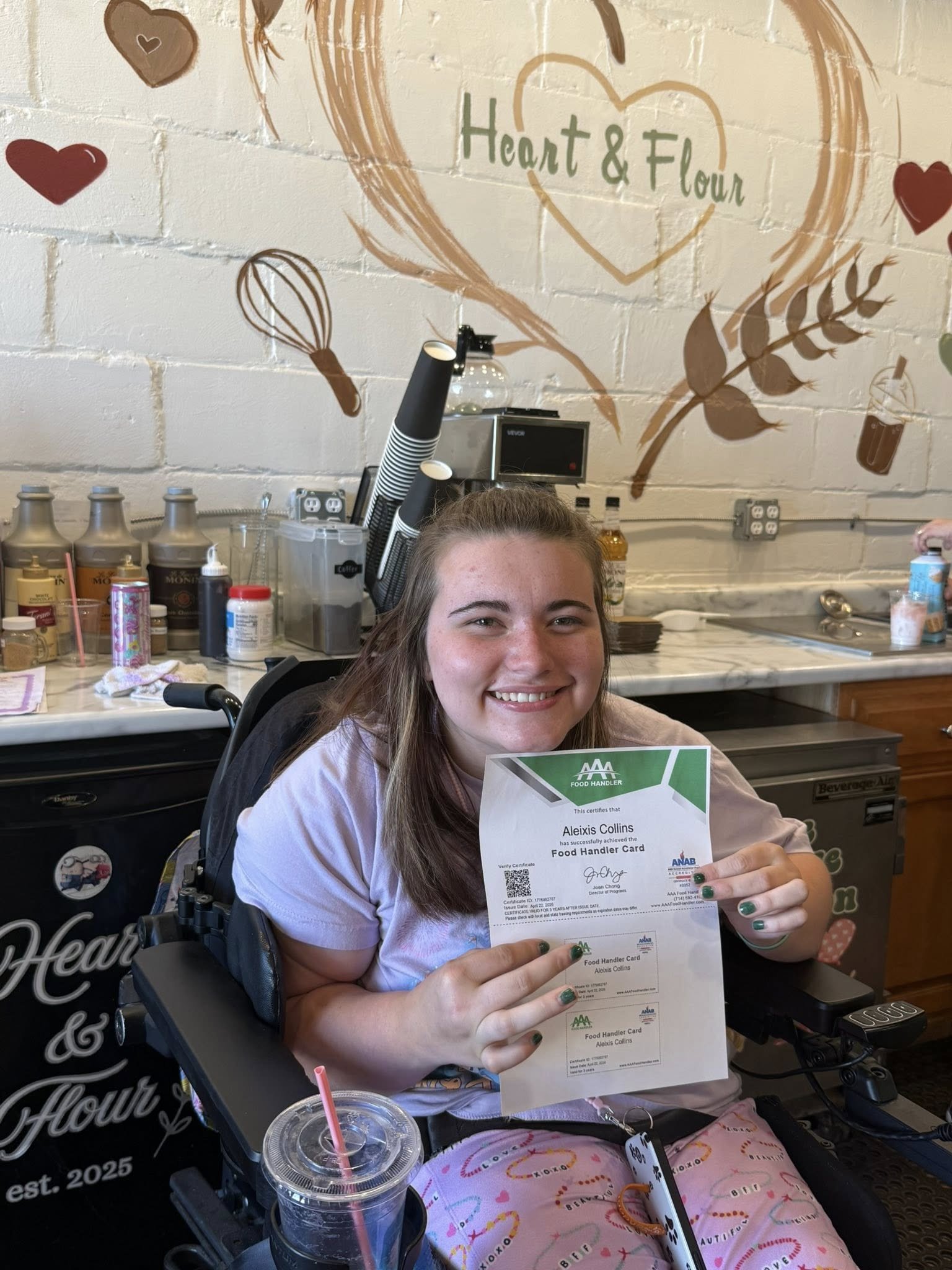 A young woman in a wheelchair smiling and holding a certificate that says she has successfully achieved the Food Handler Card.